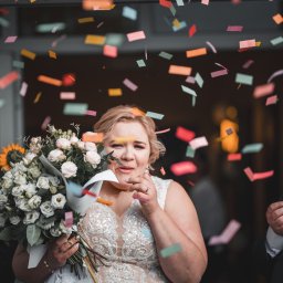 Michał Wawerek Fotografia - Panna młoda w koronkowej sukni trzyma bukiet z białych róż i słonecznika, otoczona konfetti. Obok niej mężczyźni w garniturach. Ujęcie z uroczystości ślubnej.