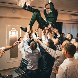 Michał Wawerek Fotografia - Radosny moment weselny: grupa mężczyzn podrzuca w górę uśmiechniętego pana młodego w zielonym garniturze, w tle wiwatujący goście i DJ z mikrofonem.