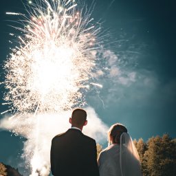 Michał Wawerek Fotografia - Para młoda wpatruje się w fajerwerki na tle nocnego nieba, stojąc na drewnianym pomoście. Widok z tyłu, suknia ślubna z welonem, garnitur.