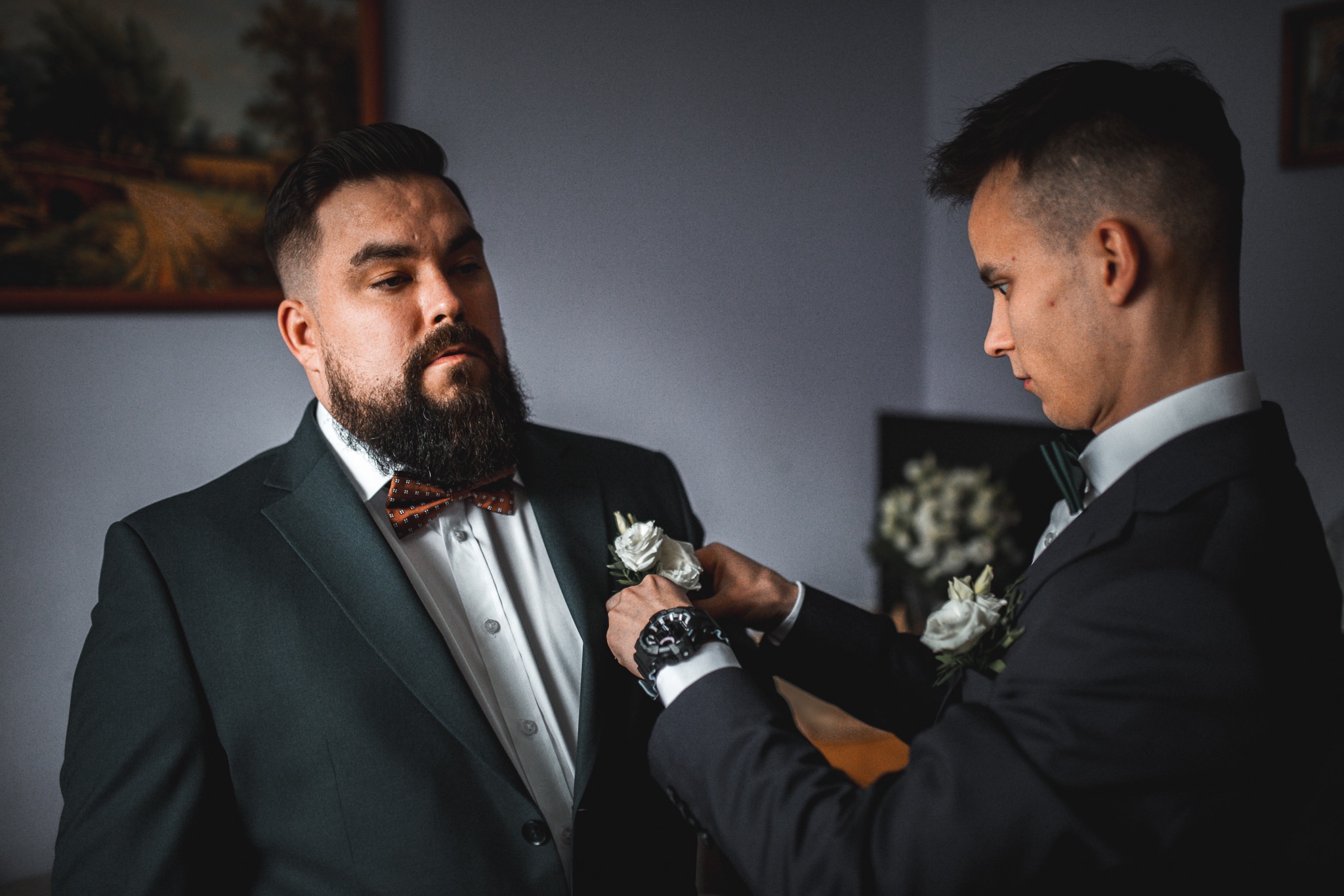 Mężczyzna w ciemnym garniturze z muszką, któremu poprawiana jest butonierka z białych róż przez drugiego mężczyznę, przygotowania do ślubu w stonowanej kolorystyce.