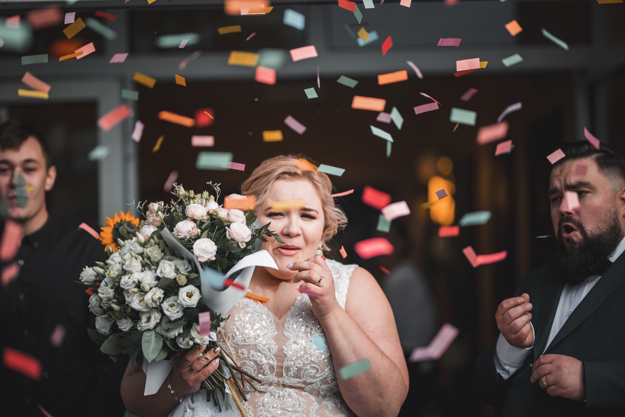 Panna młoda w koronkowej sukni trzyma bukiet z białych róż i słonecznika, otoczona konfetti. Obok niej mężczyźni w garniturach. Ujęcie z uroczystości ślubnej.