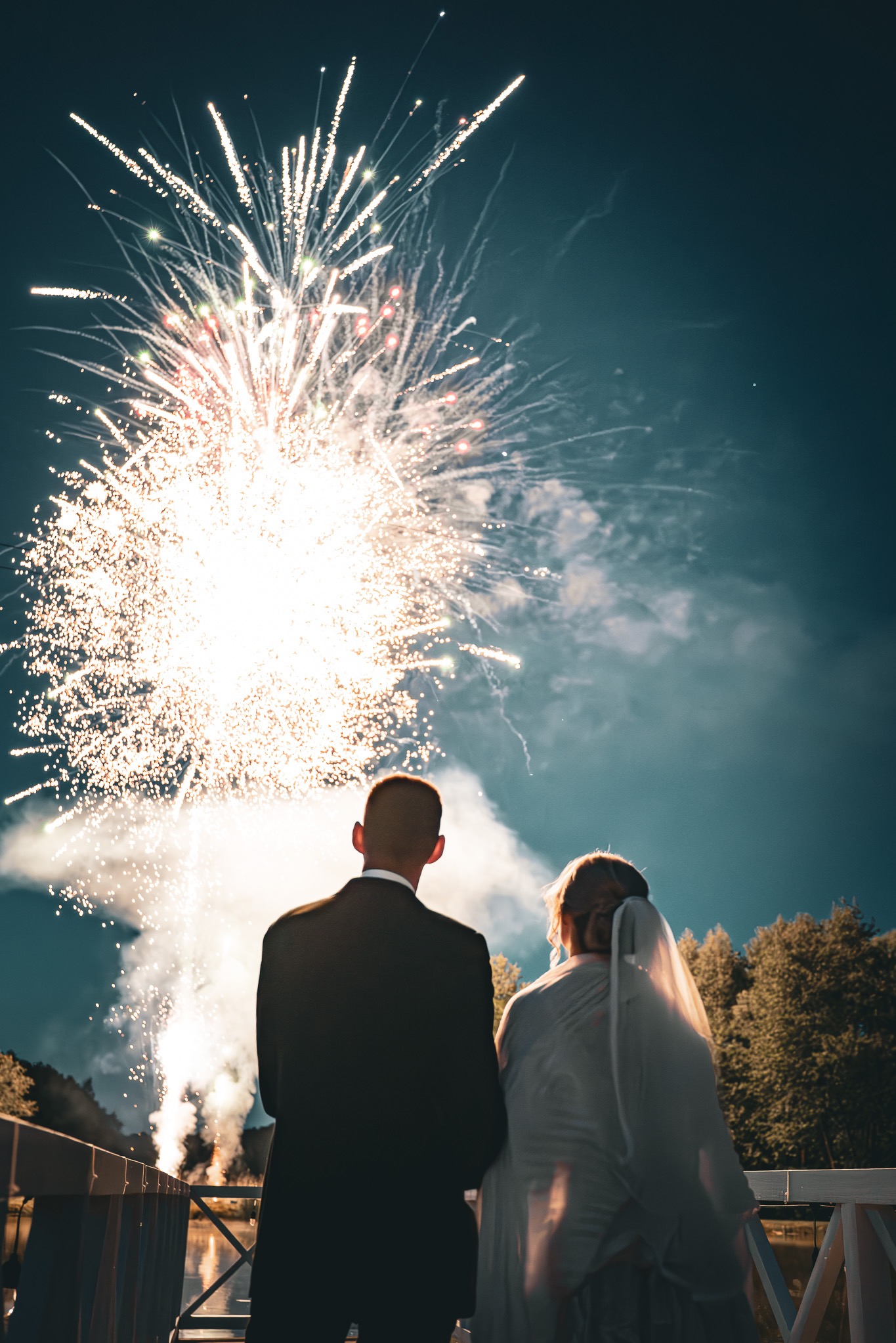 Para młoda wpatruje się w fajerwerki na tle nocnego nieba, stojąc na drewnianym pomoście. Widok z tyłu, suknia ślubna z welonem, garnitur.