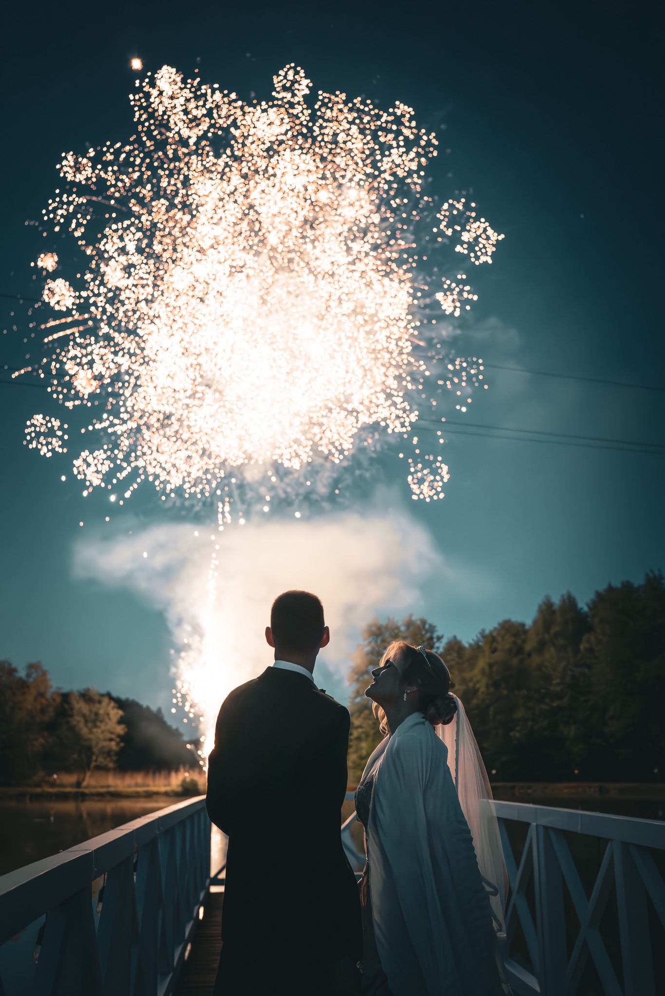 Para młoda wpatruje się w fajerwerki na tle nocnego nieba, stojąc na białym moście. Romantyczna sceneria ślubna z delikatnym oświetleniem.