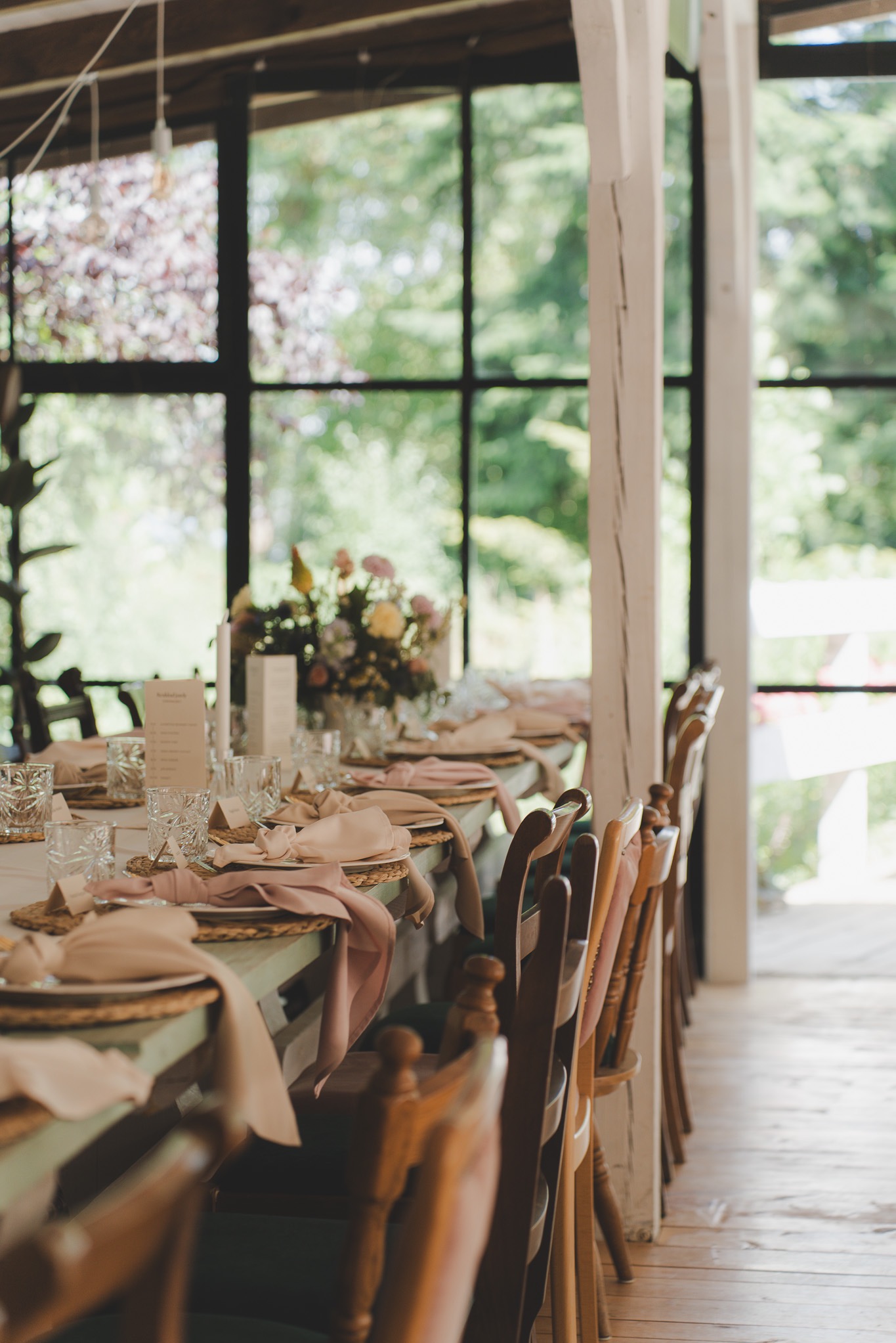 Uroczysty stół z dekoracjami kwiatowymi i elegancką zastawą, przygotowany na przyjęcie w jasnym wnętrzu z widokiem na zieleń.
