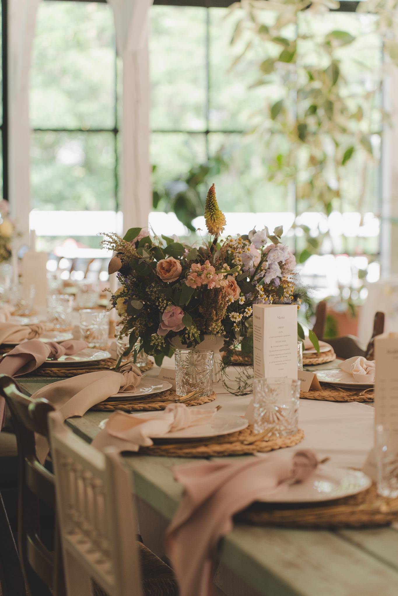 Uroczysty stół z kwiatową kompozycją, nakryty dla gości z menu i dekoracyjnymi szklankami, w jasnym wnętrzu z oknami. Pastelowe kolory, elegancki wystrój.