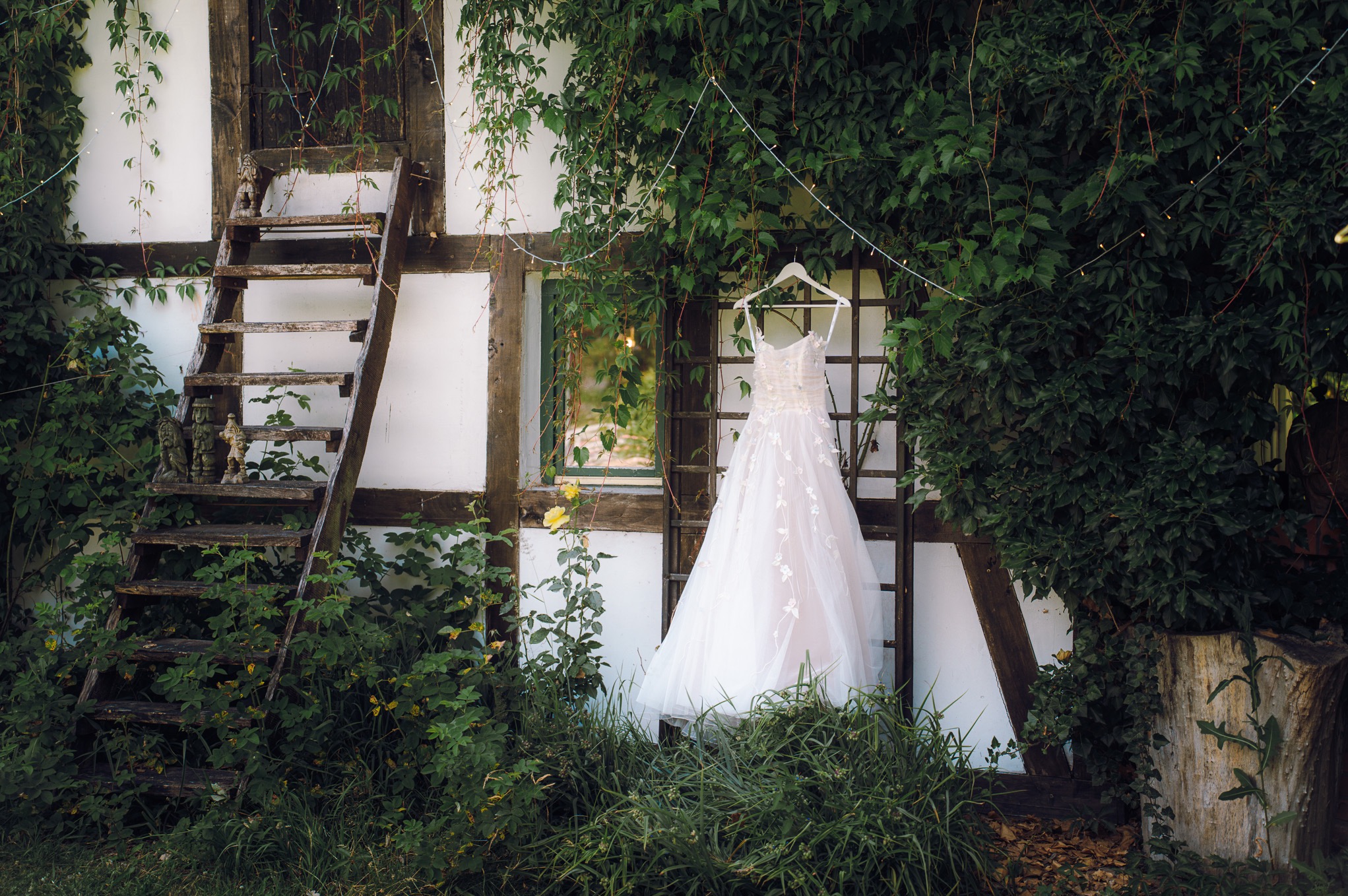 Suknia ślubna na wieszaku, wisząca na tle ściany porośniętej bluszczem, obok drewniana drabina i okno. Romantyczna sceneria.