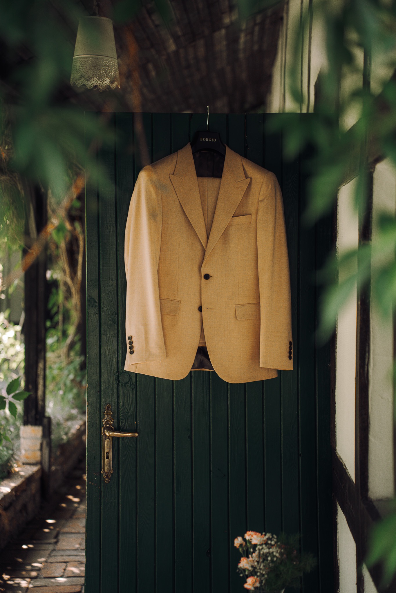 Beżowy garnitur męski marki Borgio zawieszony na zielonych drzwiach, z widocznymi detalami jak guziki i faktura materiału, w otoczeniu zieleni i kwiatów.