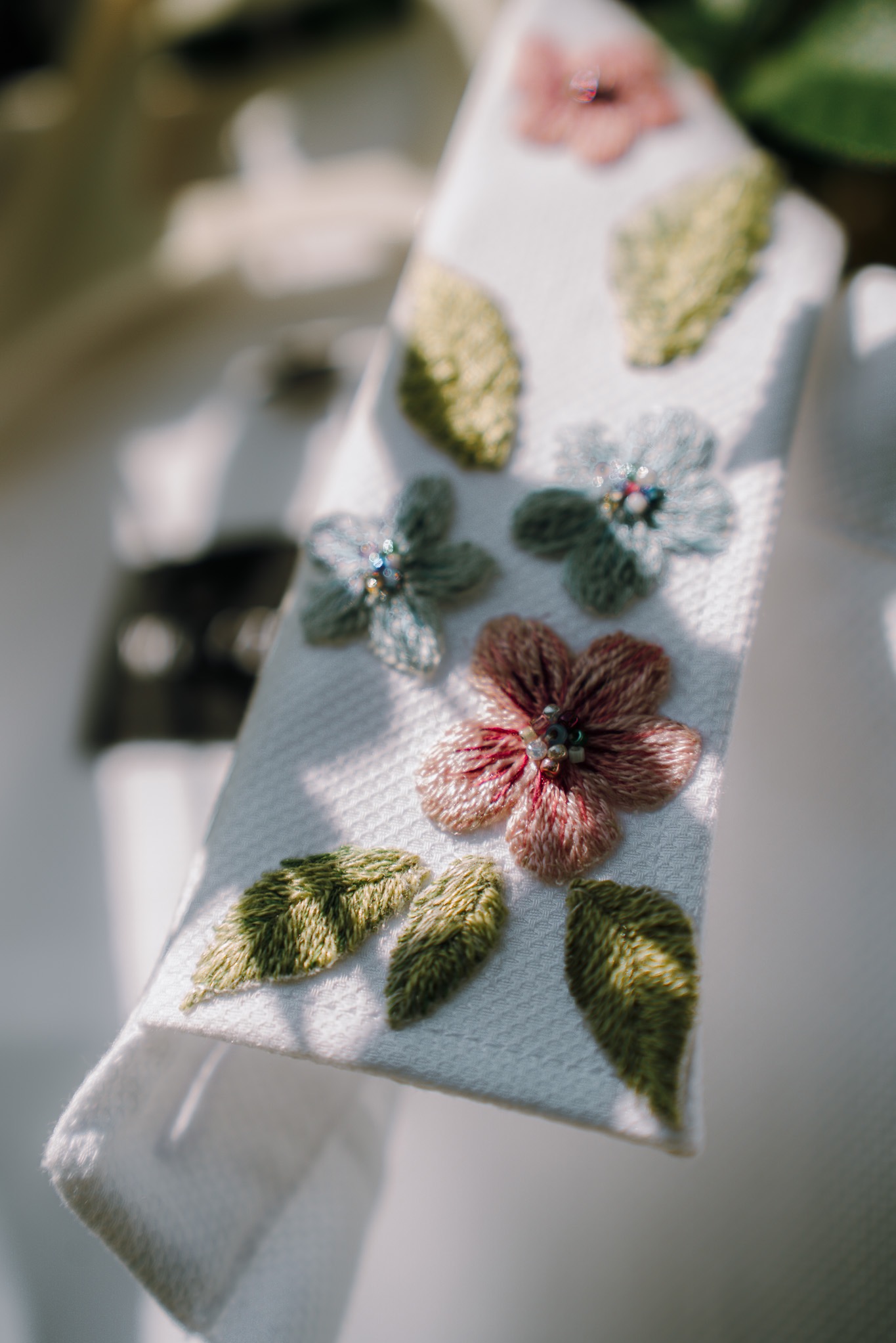 Biała tkanina zdobiona haftowanymi kwiatami i listkami w pastelowych odcieniach, z detalami z koralików, uchwycona w naturalnym świetle.