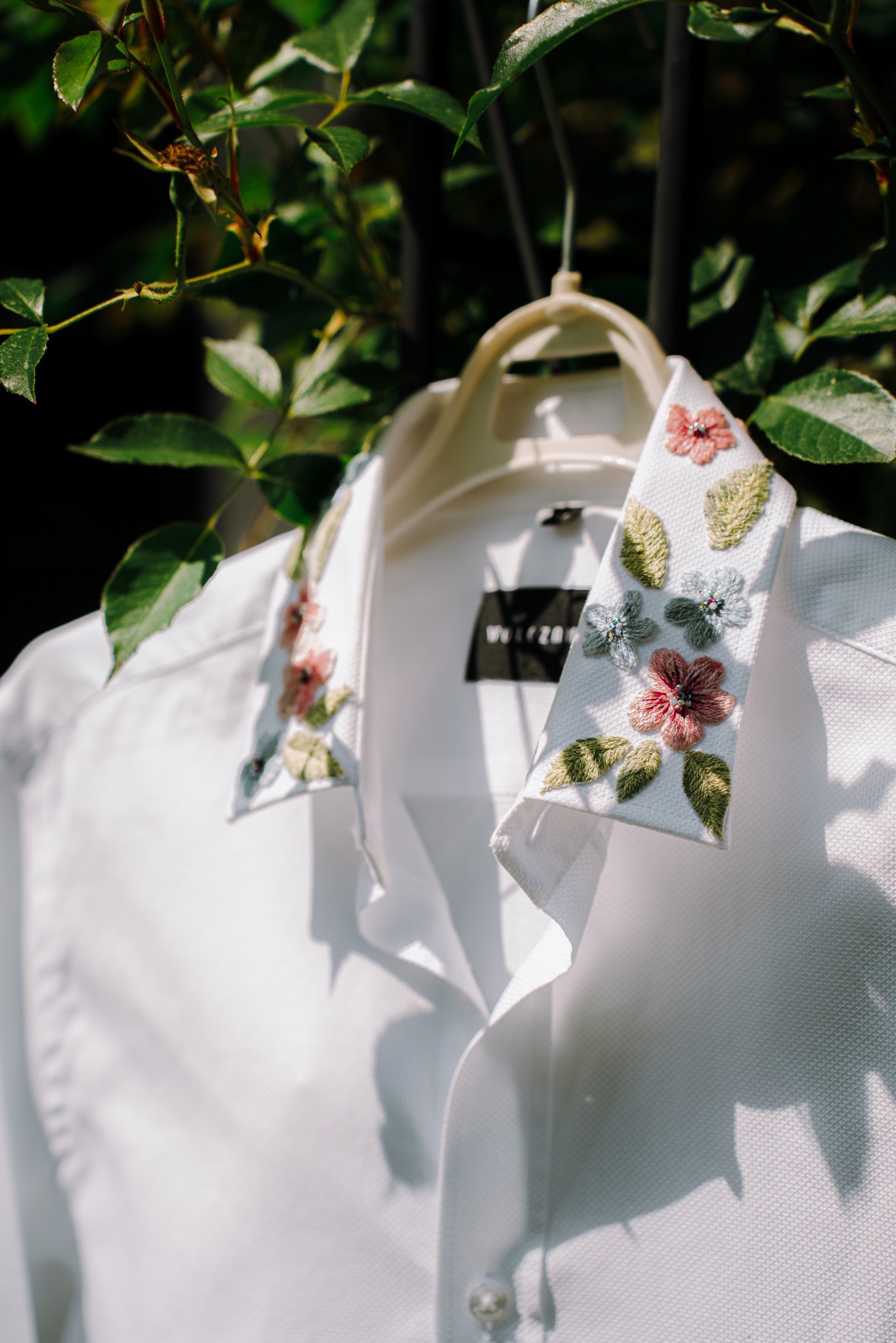 Biała koszula z haftowanymi kwiatami na kołnierzyku, wisząca na wieszaku na tle zielonych liści. Delikatne światło podkreśla detale haftu.