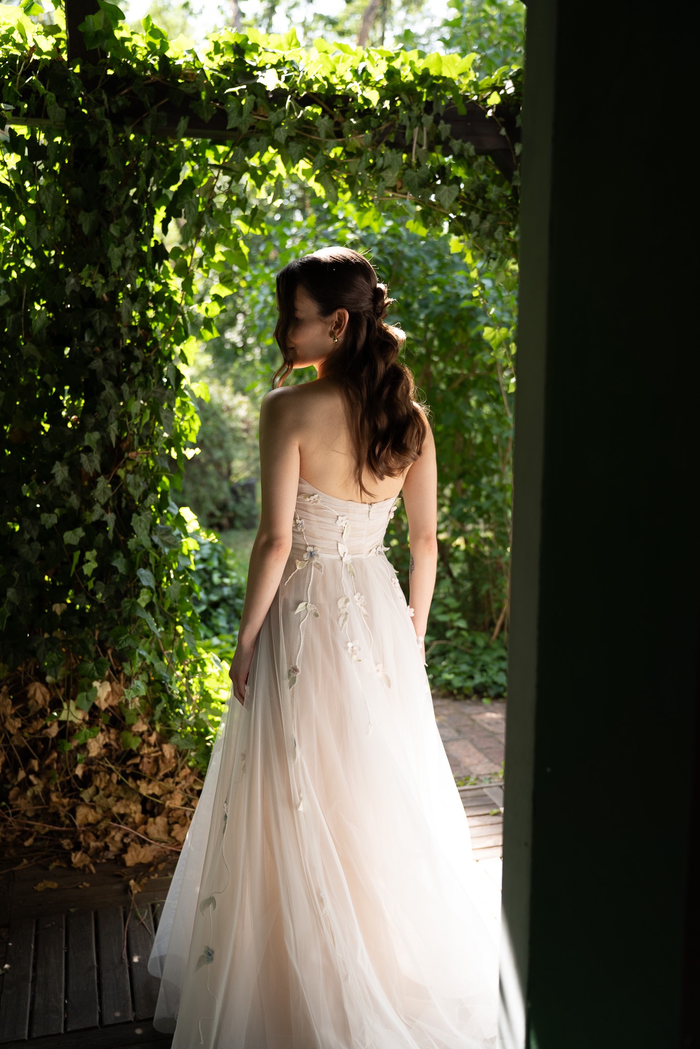 Kobieta w zwiewnej sukni, odwrócona tyłem, na tle zielonej roślinności i drewnianej konstrukcji. Delikatne światło podkreśla detale sukni i fryzury.