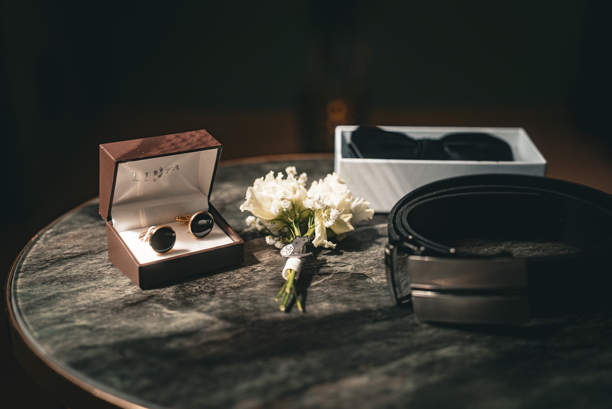 Eleganckie detale ślubne: spinki do mankietów w pudełku, butonierka z przypinką kota, pasek i mucha na marmurowym stoliku w stonowanej kolorystyce.