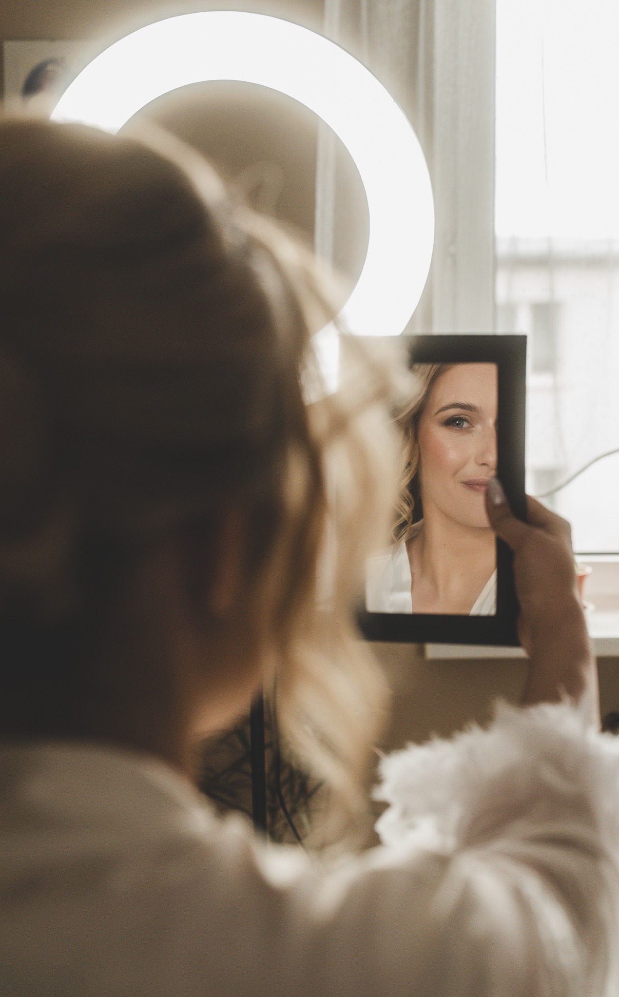 Portret kobiety z makijażem, widziana w lustrze trzymanym w dłoni, z lampą pierścieniową w tle. Ujęcie zza ramienia, skupione na odbiciu.