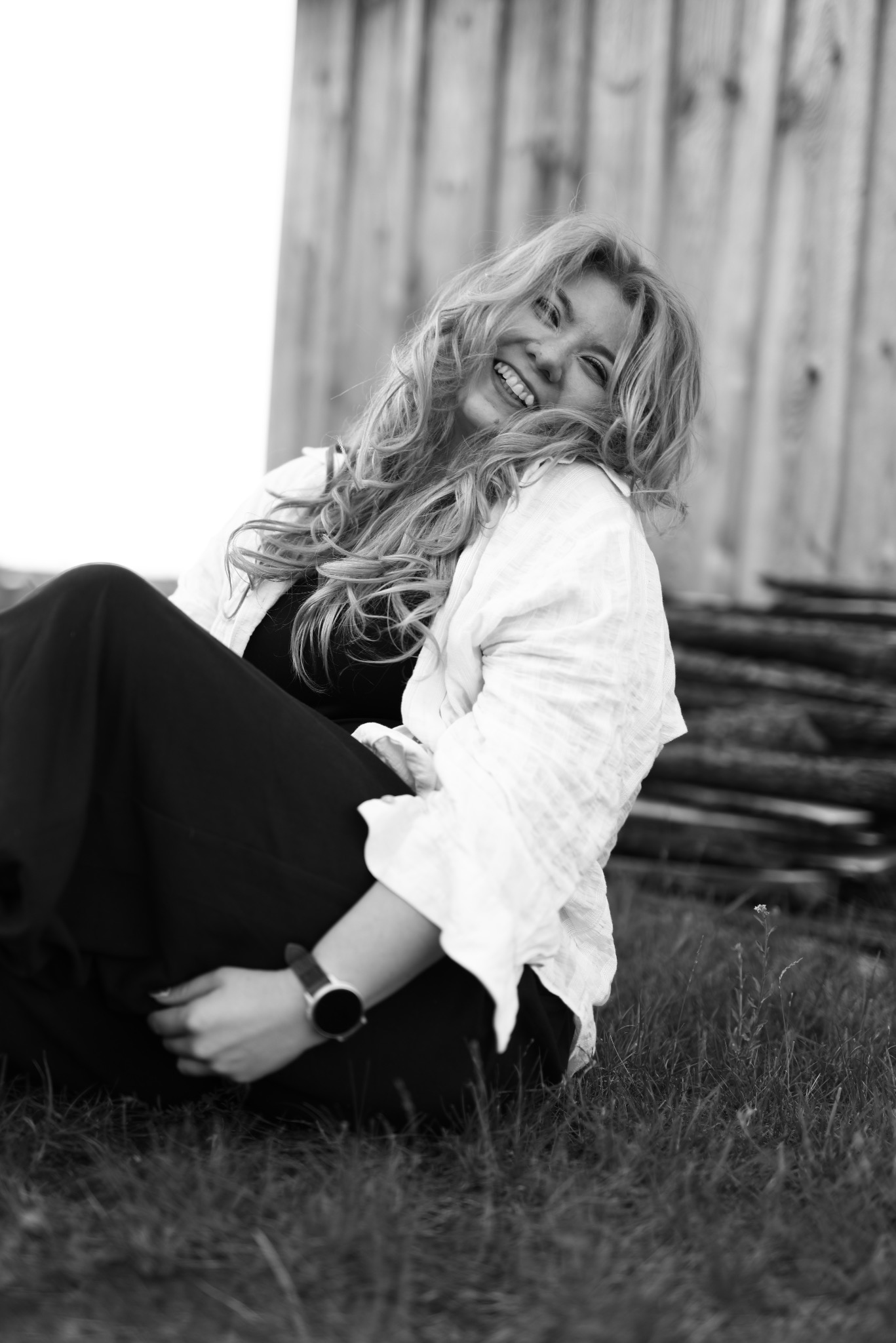 Uśmiechnięta blondynka w białej koszuli siedzi na trawie, w tle drewniana ściana. Zdjęcie czarno-białe, z bliska, z widocznym zegarkiem na ręce.