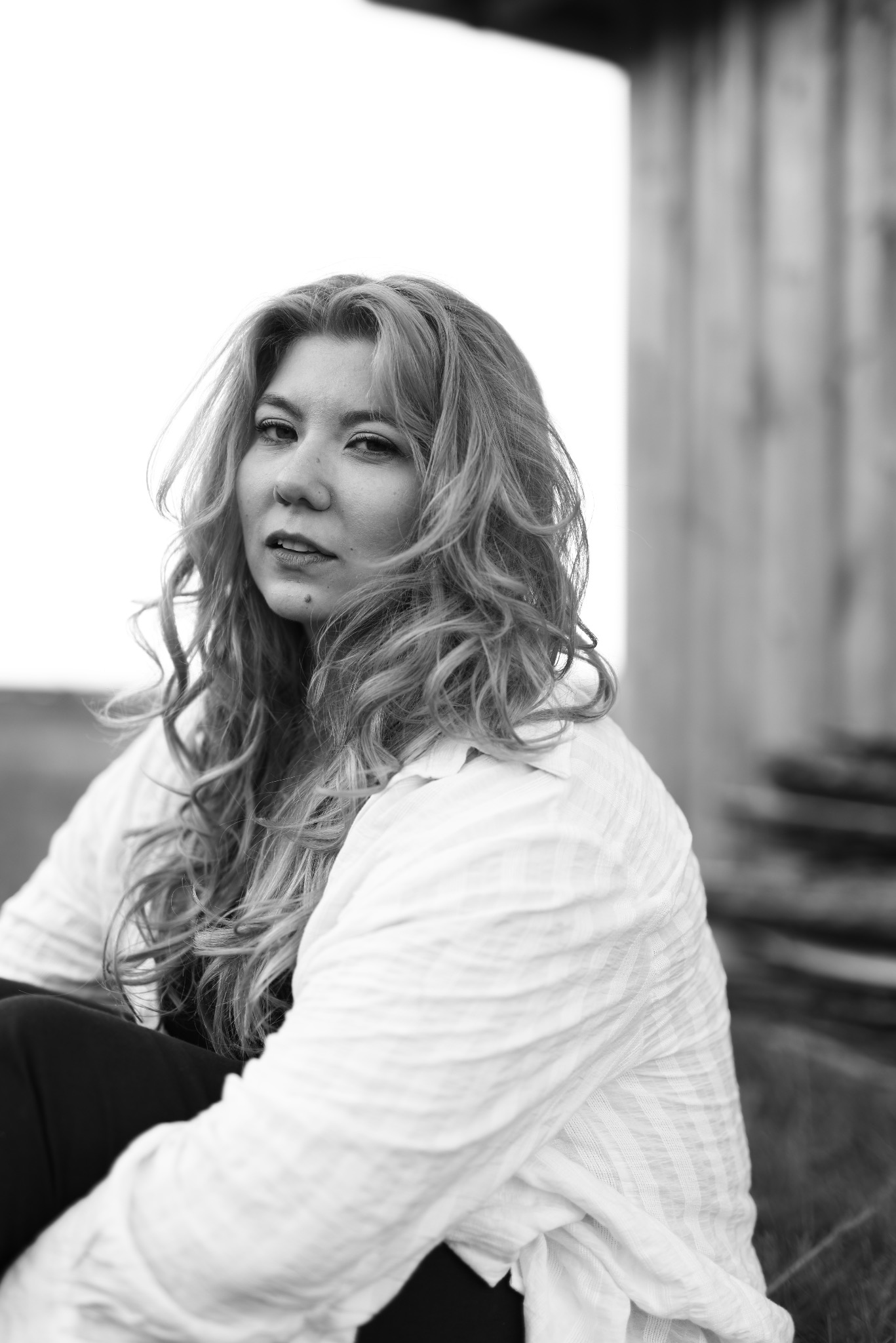 Portret kobiety o kręconych włosach w białej koszuli na tle jasnego nieba i drewnianej konstrukcji. Zdjęcie czarno-białe, z delikatnym oświetleniem.