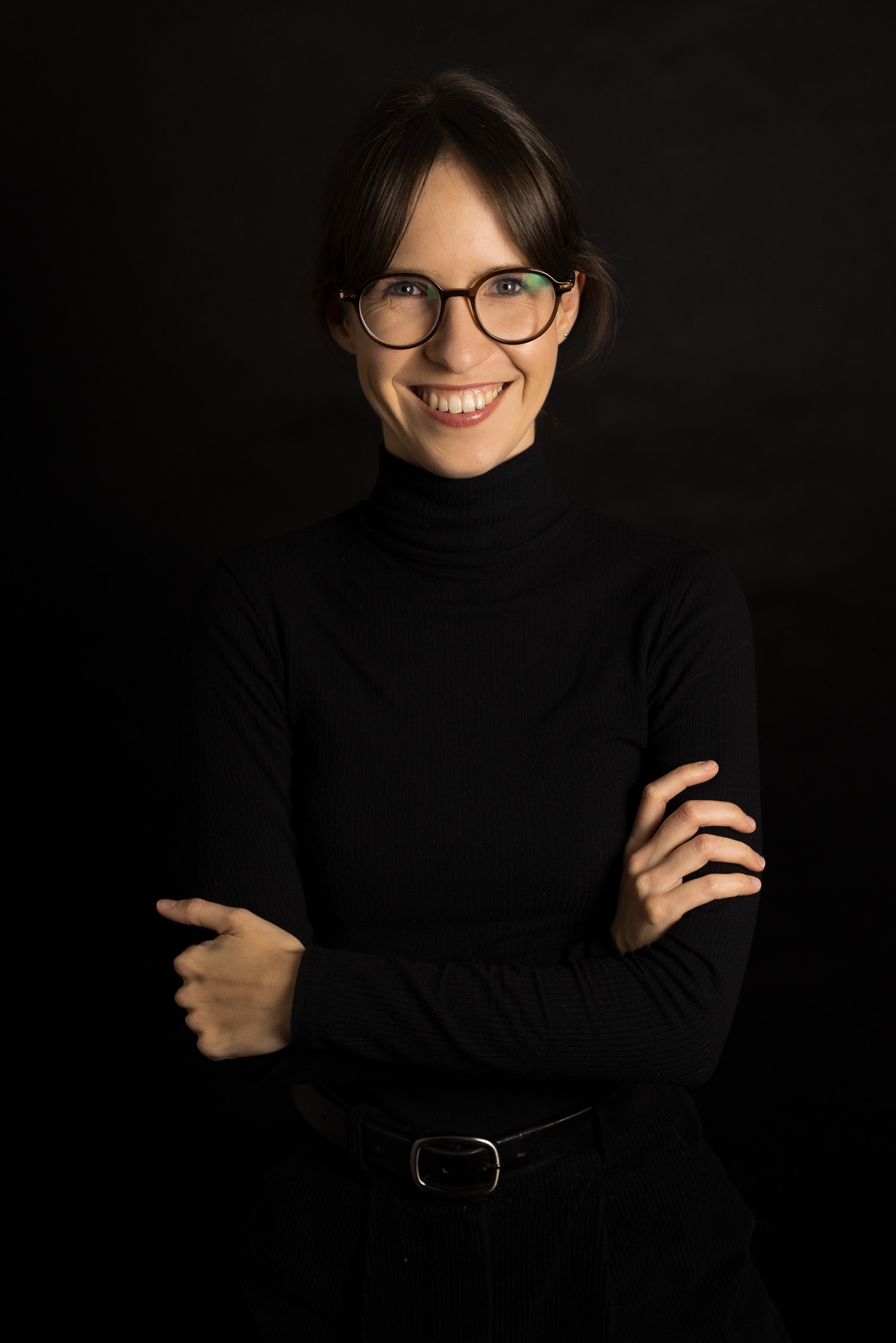 Portret uśmiechniętej kobiety w okularach, ubrana w czarny golf, z rękami skrzyżowanymi na piersiach na jednolitym, ciemnym tle. Profesjonalny wizerunek.