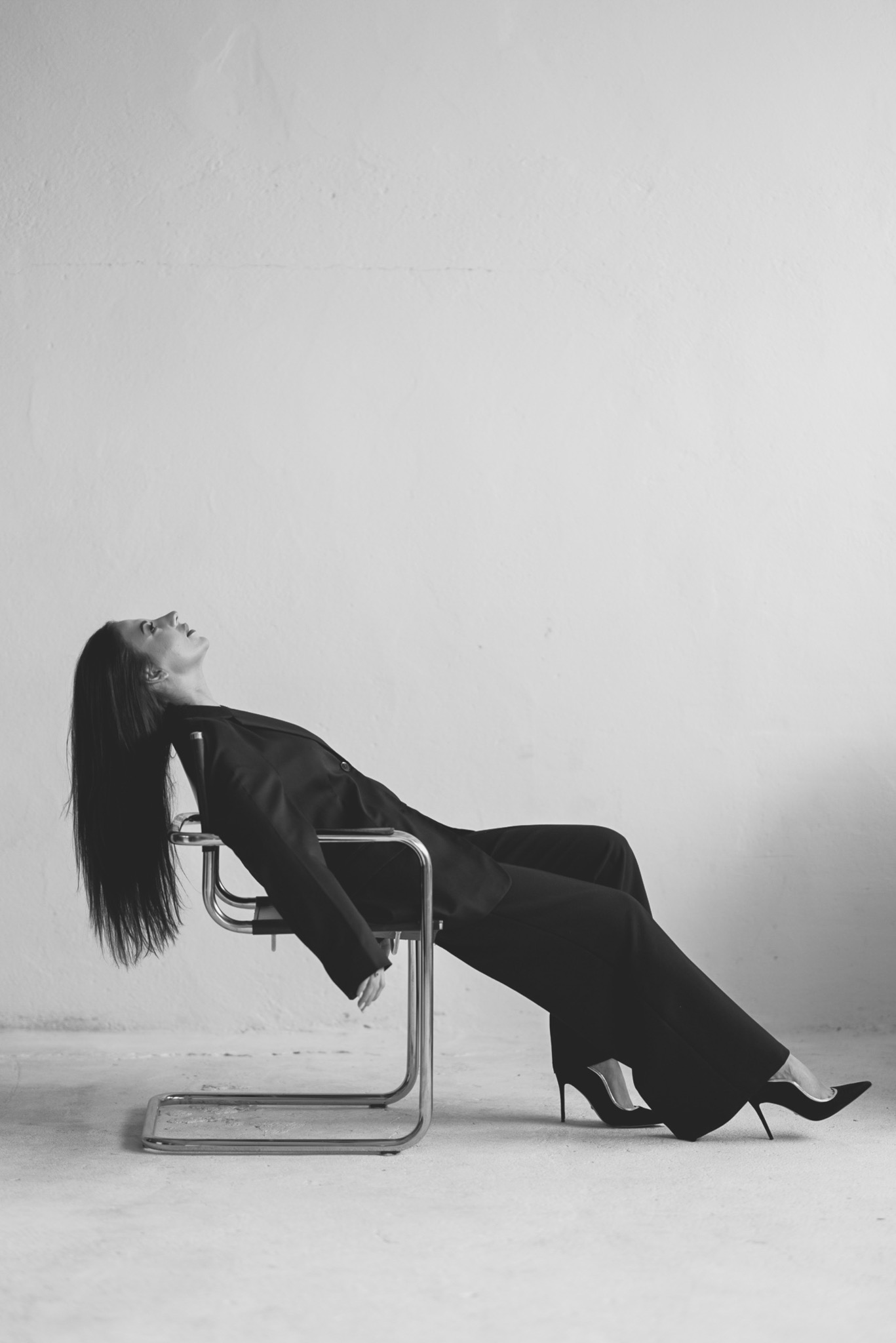 Czarno-białe zdjęcie kobiety w garniturze i szpilkach, leżącej na krześle w minimalistycznym wnętrzu. Długie włosy opadają swobodnie, tworząc kontrast z jasnym tłem.