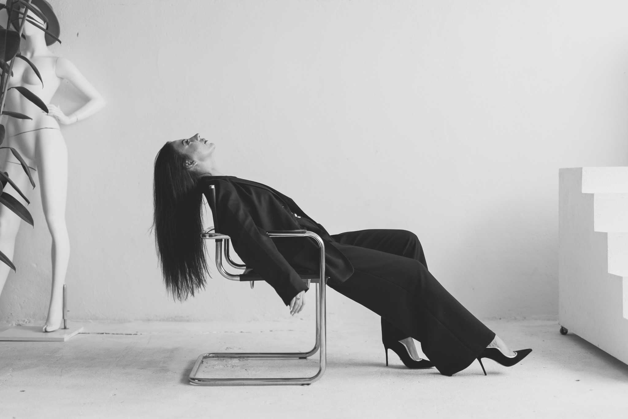 Czarno-białe zdjęcie kobiety w garniturze, leżącej na krześle w minimalistycznym wnętrzu z manekinem i rośliną w tle. Ujęcie z niskiego kąta, podkreślające długie włosy modelki.