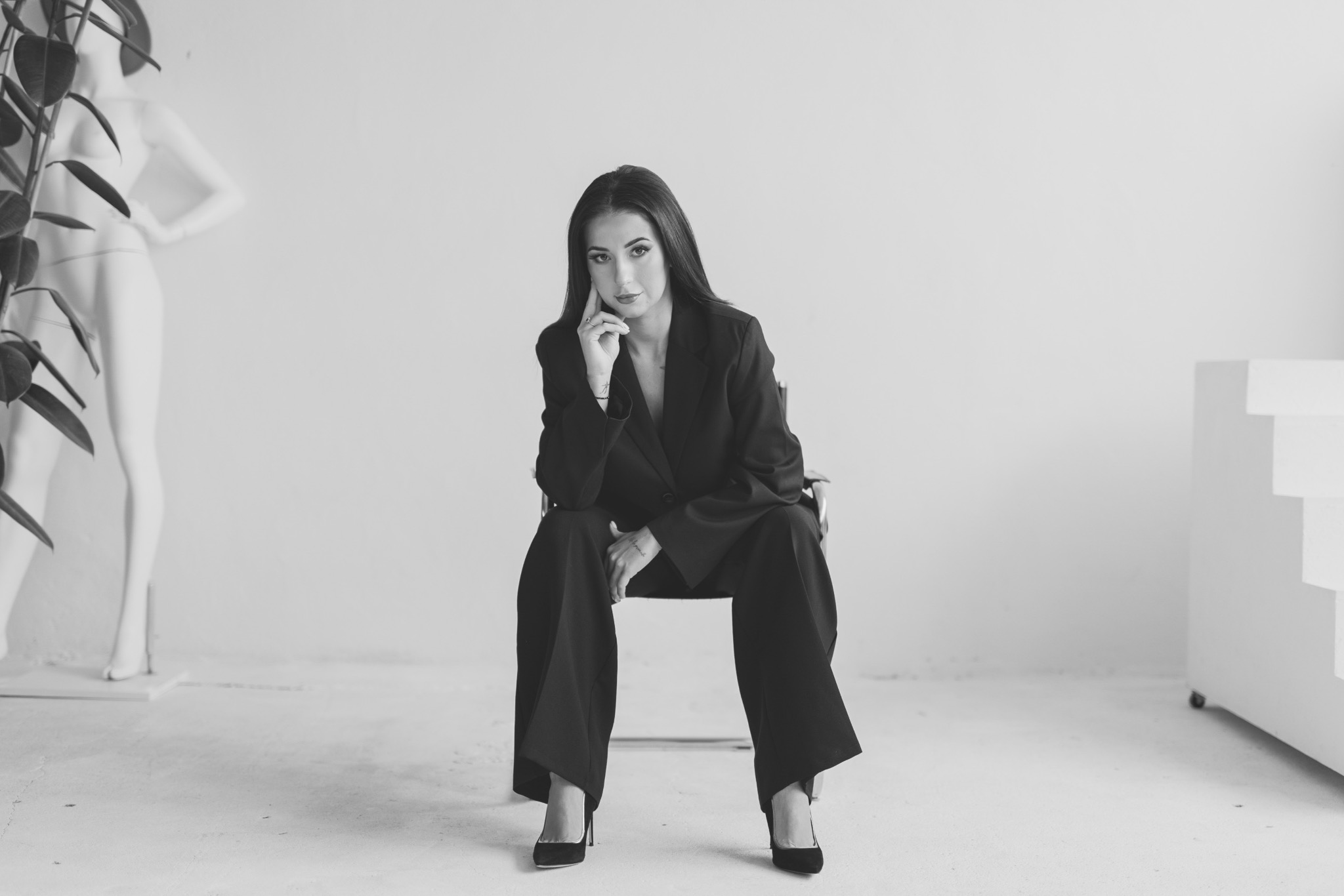 Monochromatyczne ujęcie kobiety w czarnym garniturze, siedzącej na krześle w minimalistycznym studio z rośliną i manekinem w tle. Uwagę przykuwa jej wyrazisty makijaż.