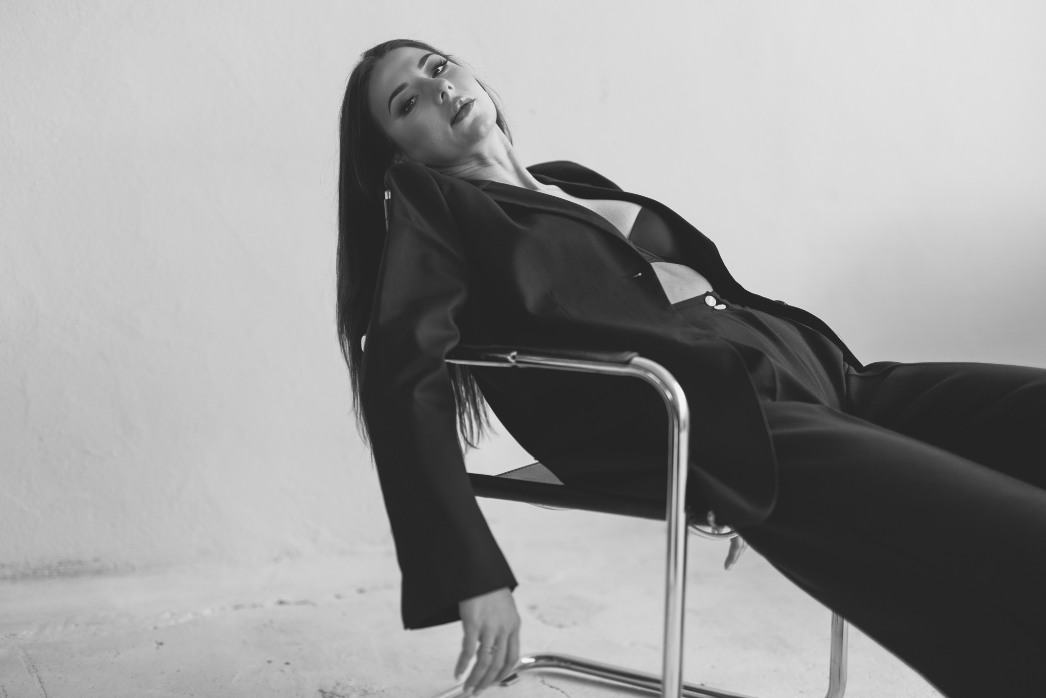 Monochromatyczne ujęcie kobiety w czarnym garniturze, siedzącej na krześle na jasnym tle. Uwagę przyciąga jej nonszalancka poza i wyrazisty makijaż.