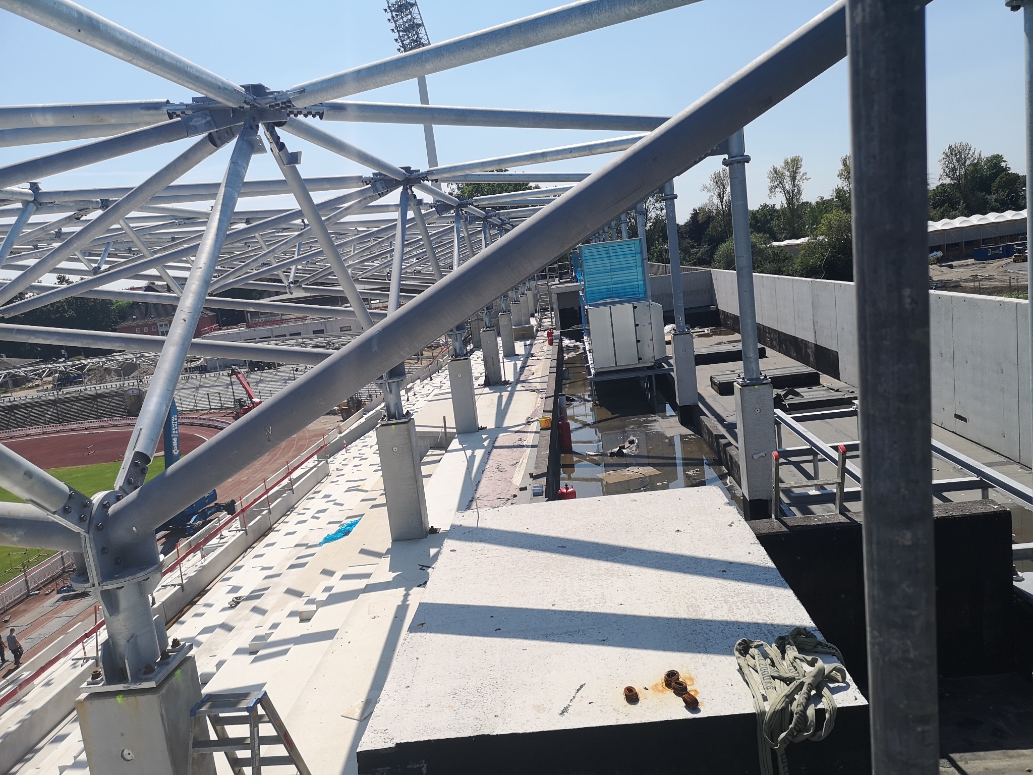Stalowa konstrukcja dachu stadionu w trakcie budowy. Widoczne elementy wentylacji, instalacje i drabina. Jasne niebo w tle.