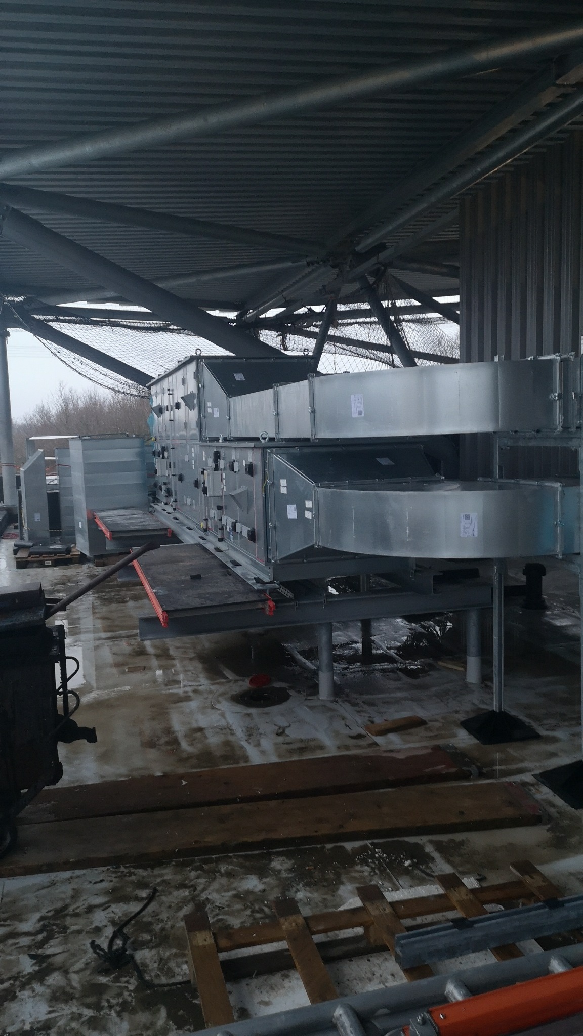 Szare, metalowe moduły wentylacyjne, prawdopodobnie klimatyzacyjne, ustawione na platformie pod dachem budynku w trakcie instalacji. Widoczne elementy konstrukcyjne dachu.