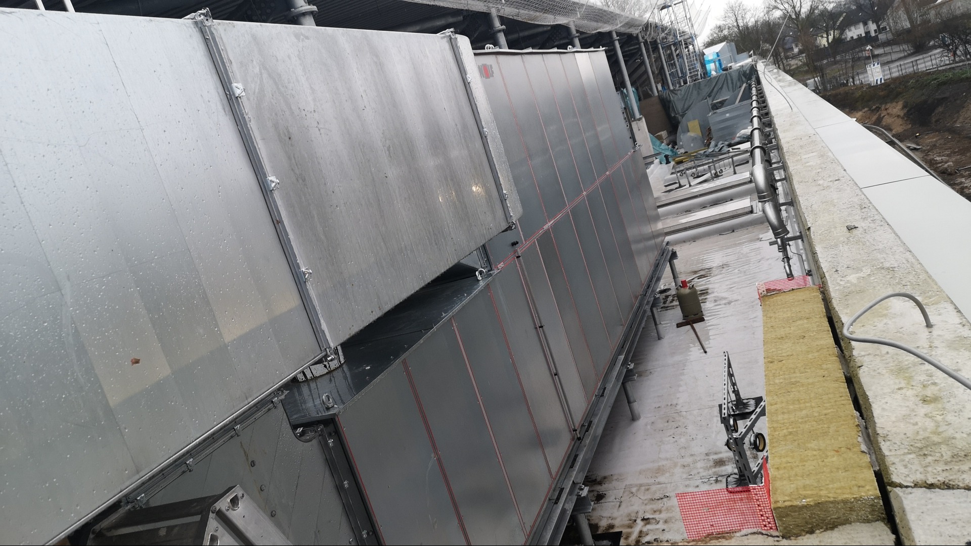 Widok z góry na zainstalowany system wentylacyjny z rurami na dachu budynku w trakcie montażu, z widoczną izolacją z wełny mineralnej i butlą gazową.