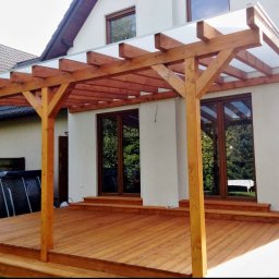 Zakład Stolarski StyloveWood Maciej Grabowski - Drewniana pergola z zadaszeniem z poliwęglanu, przylegająca do budynku z dwoma wejściami. Taras z desek i basen w tle. Naturalne światło.