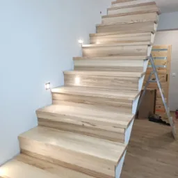 Jasne, drewniane schody z subtelnym oświetleniem punktowym wzdłuż ściany, widoczne z dołu. W tle drabina oparta o ścianę i drzwi z drewnianymi panelami.