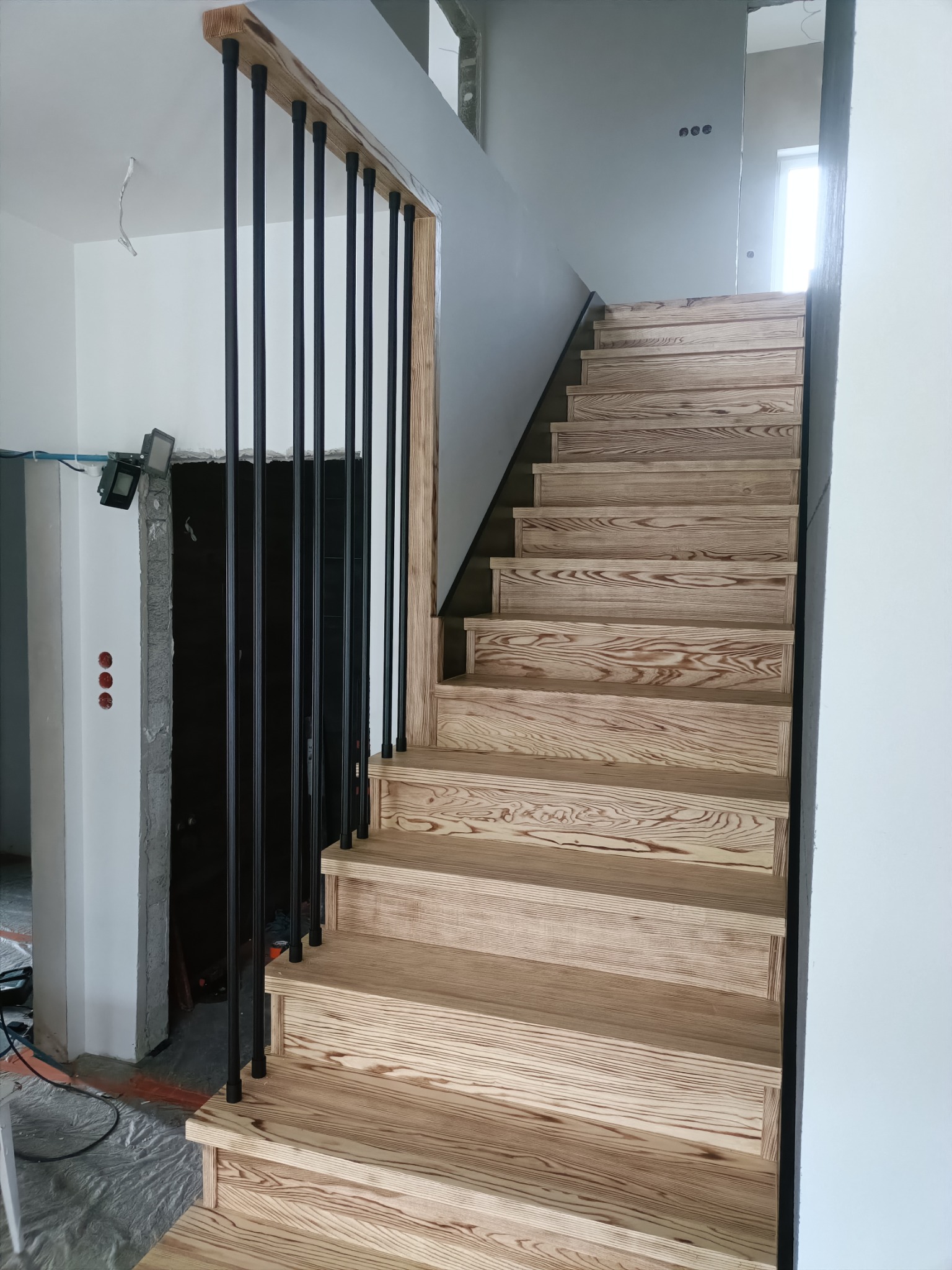 Jasne, drewniane schody z czarną, metalową balustradą w surowym wnętrzu. Widoczne ślady prac wykończeniowych, podkreślające etap budowy.