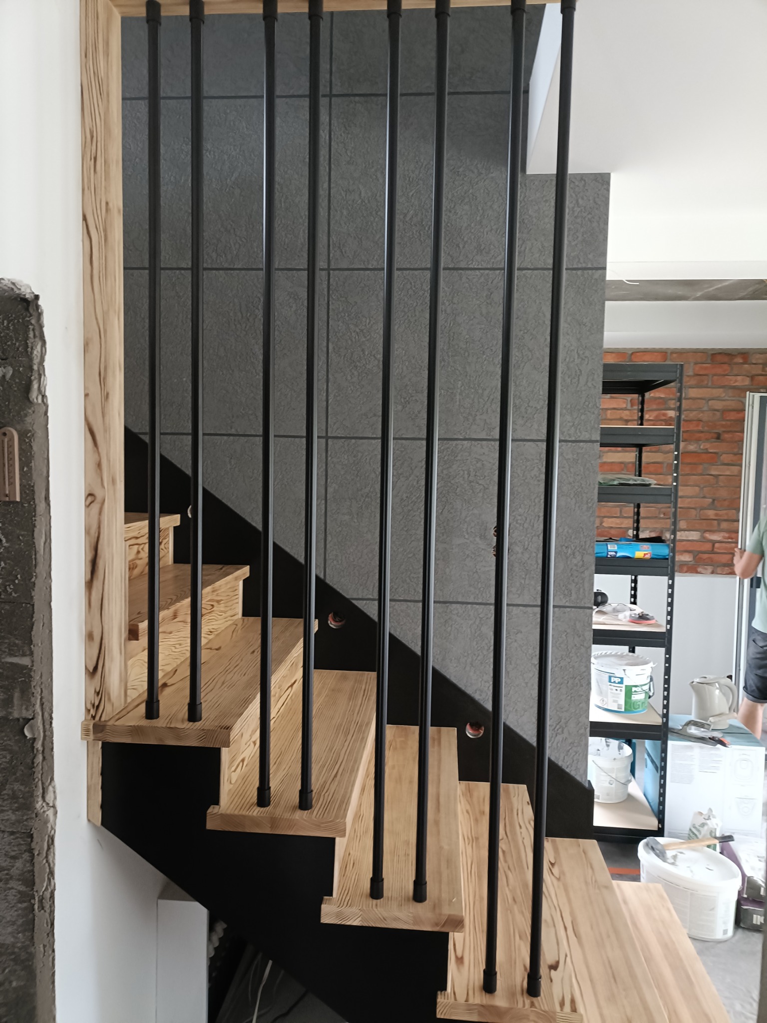 Nowoczesne schody z jasnego drewna z czarnymi, metalowymi balustradami na tle szarej ściany z płytek. Widoczne fragmenty pomieszczenia w trakcie wykończenia.