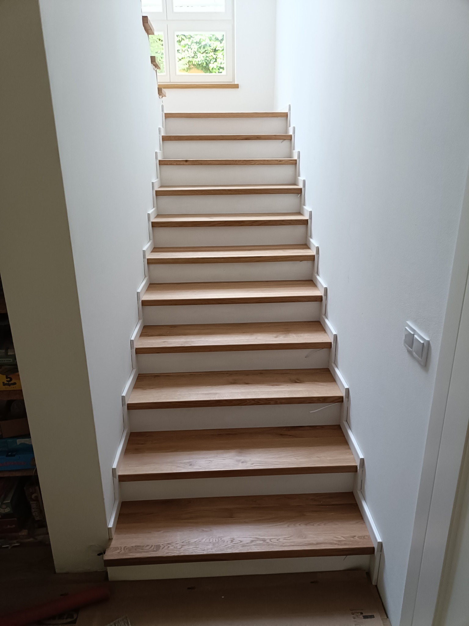 Proste, nowoczesne schody drewniane z białymi podstopnicami w jasnym wnętrzu. Naturalne drewno stopni kontrastuje z bielą ścian i minimalistycznym stylem.