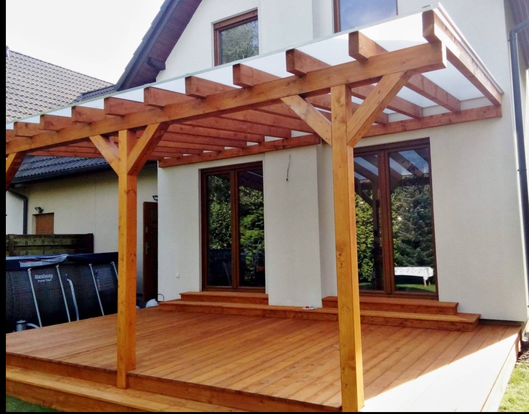 Drewniana pergola z zadaszeniem z poliwęglanu, przylegająca do budynku z dwoma wejściami. Taras z desek i basen w tle. Naturalne światło.