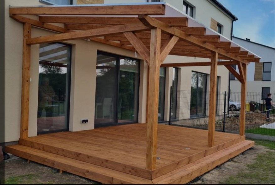 Drewniana pergola z zadaszeniem z poliwęglanu, przylegająca do nowoczesnego domu. Taras wyłożony deskami, widoczne schody i słupy konstrukcyjne.