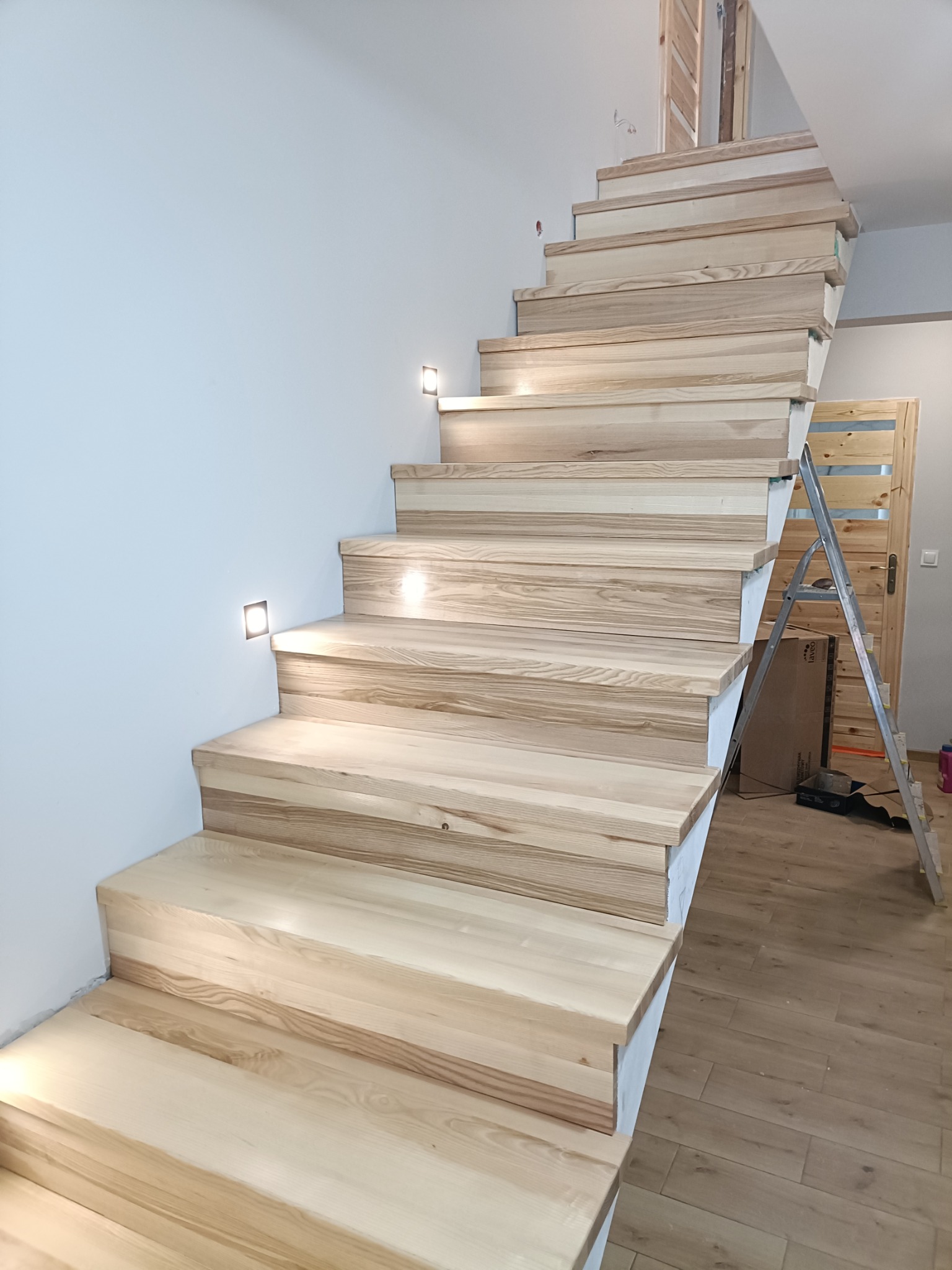 Jasne, drewniane schody z subtelnym oświetleniem punktowym wzdłuż ściany, widoczne z dołu. W tle drabina oparta o ścianę i drzwi z drewnianymi panelami.