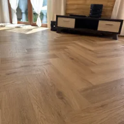 Ułożone panele podłogowe w jodełkę, z widocznym fragmentem okna z białymi zasłonami i szafką RTV z drewnianym panelem na ścianie w tle. Jasne wnętrze.