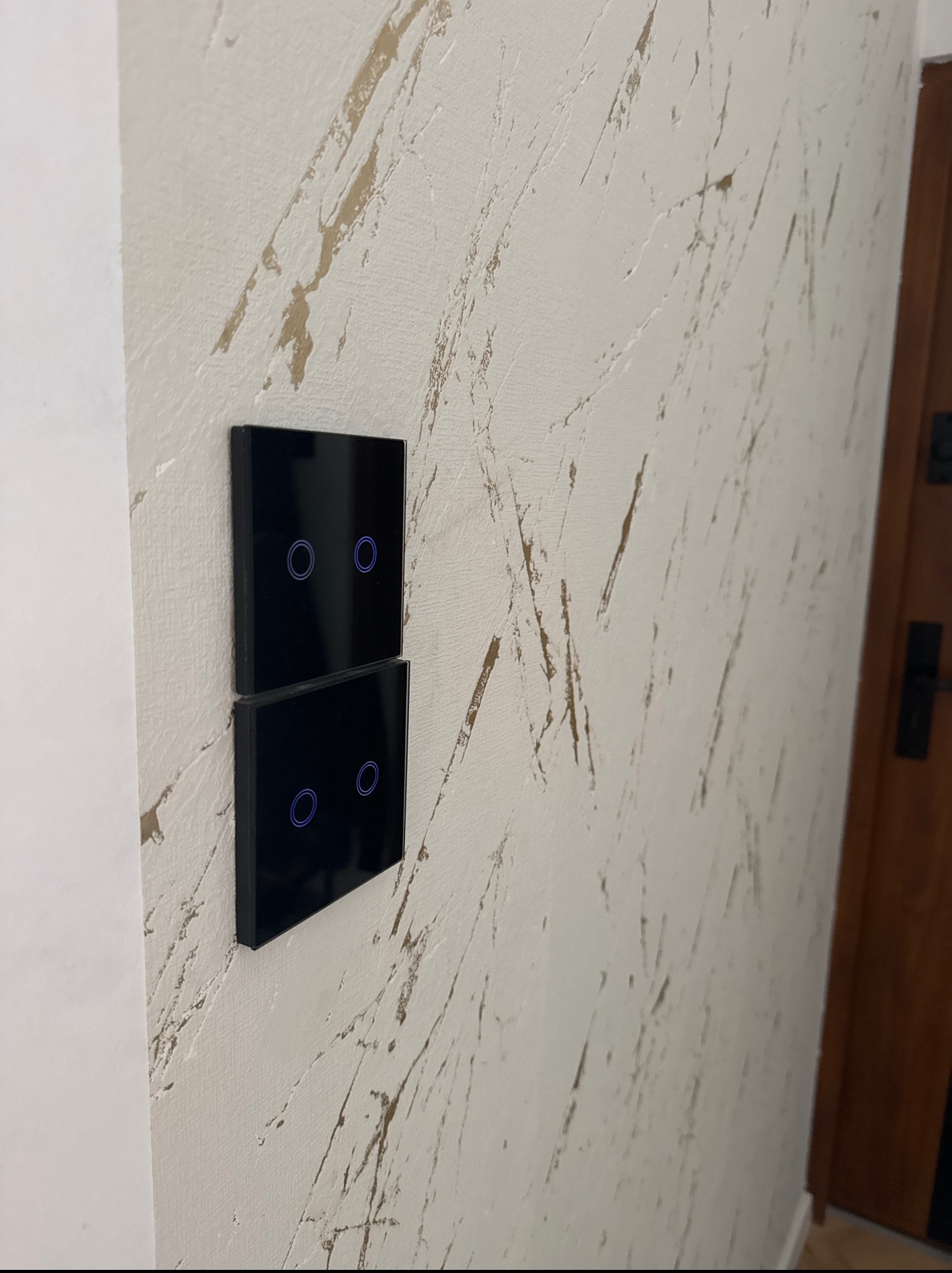 Dwa czarne, dotykowe włączniki światła zamontowane na ścianie z dekoracyjną fakturą przypominającą marmur. Widoczny fragment drzwi.