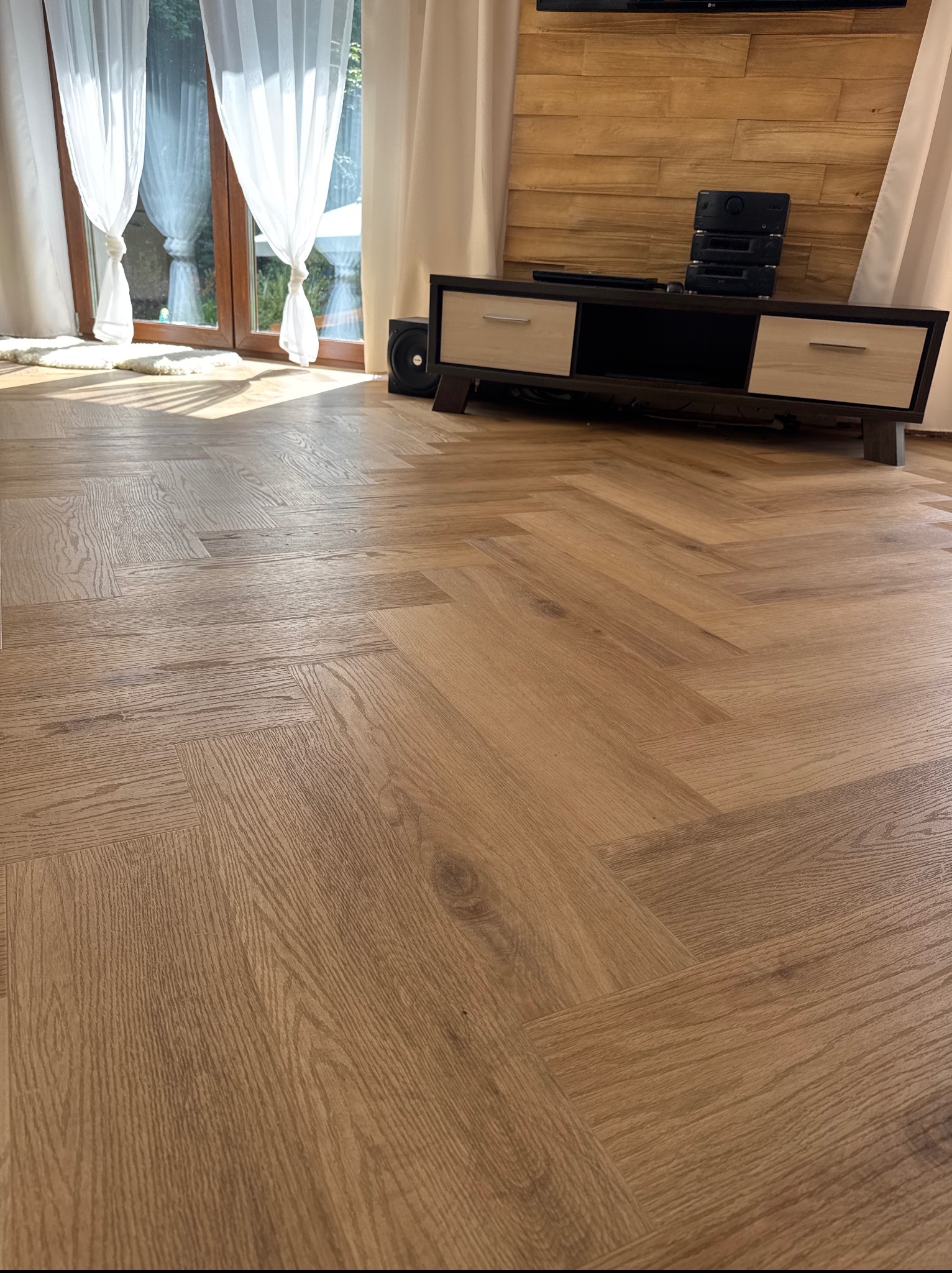 Ułożone panele podłogowe w jodełkę, z widocznym fragmentem okna z białymi zasłonami i szafką RTV z drewnianym panelem na ścianie w tle. Jasne wnętrze.