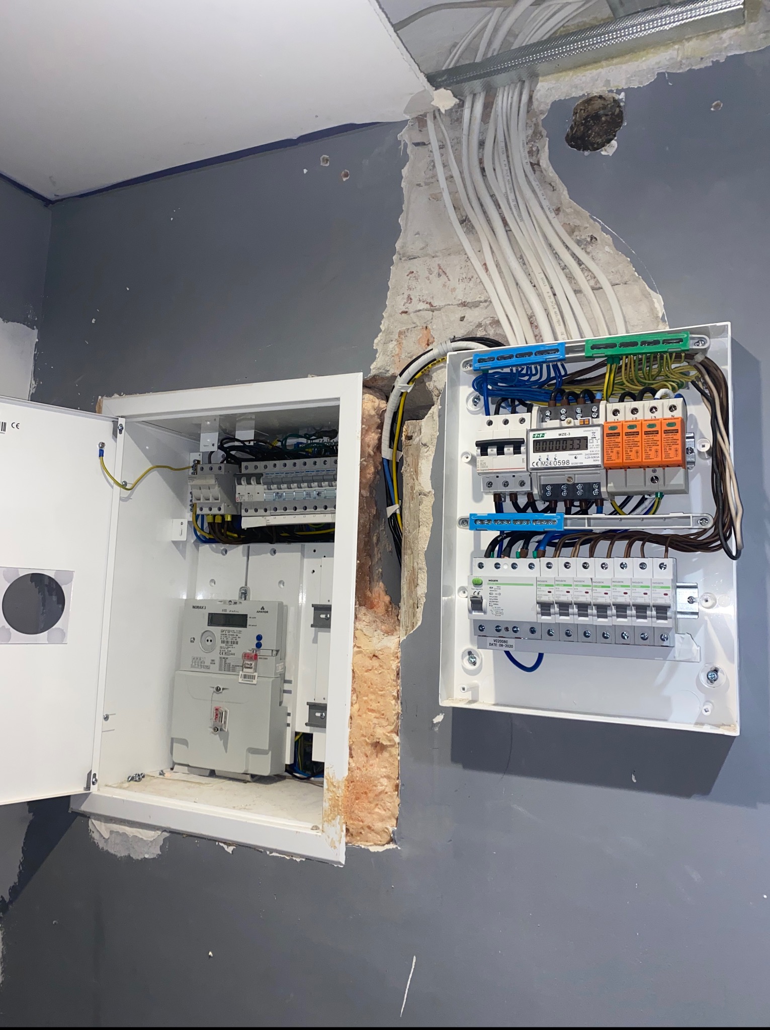 Rozdzielnia elektryczna z licznikiem, widoczne zabezpieczenia i kable. Otwarta skrzynka po lewej stronie, przewody prowadzone w listwie na suficie, ściana w szarym kolorze.