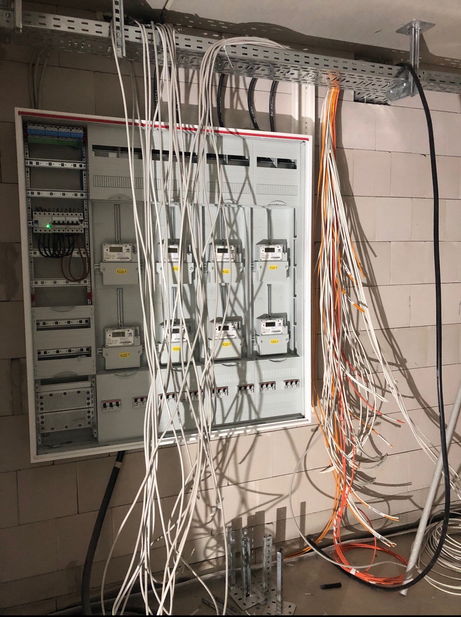 Rozdzielnica elektryczna z podłączonymi przewodami w surowym wnętrzu. Widoczne liczniki i zabezpieczenia. Przewody ułożone w korytkach kablowych pod sufitem.