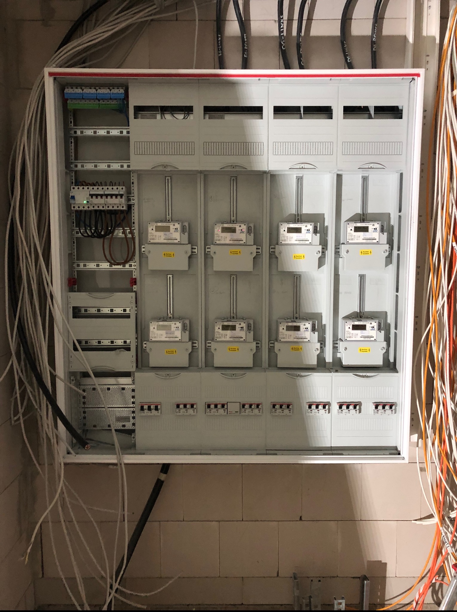 Rozdzielnia elektryczna z zamontowanymi licznikami energii, widoczne kable i zabezpieczenia. Ściana z bloczków gazobetonowych w tle. Oświetlenie sufitowe.