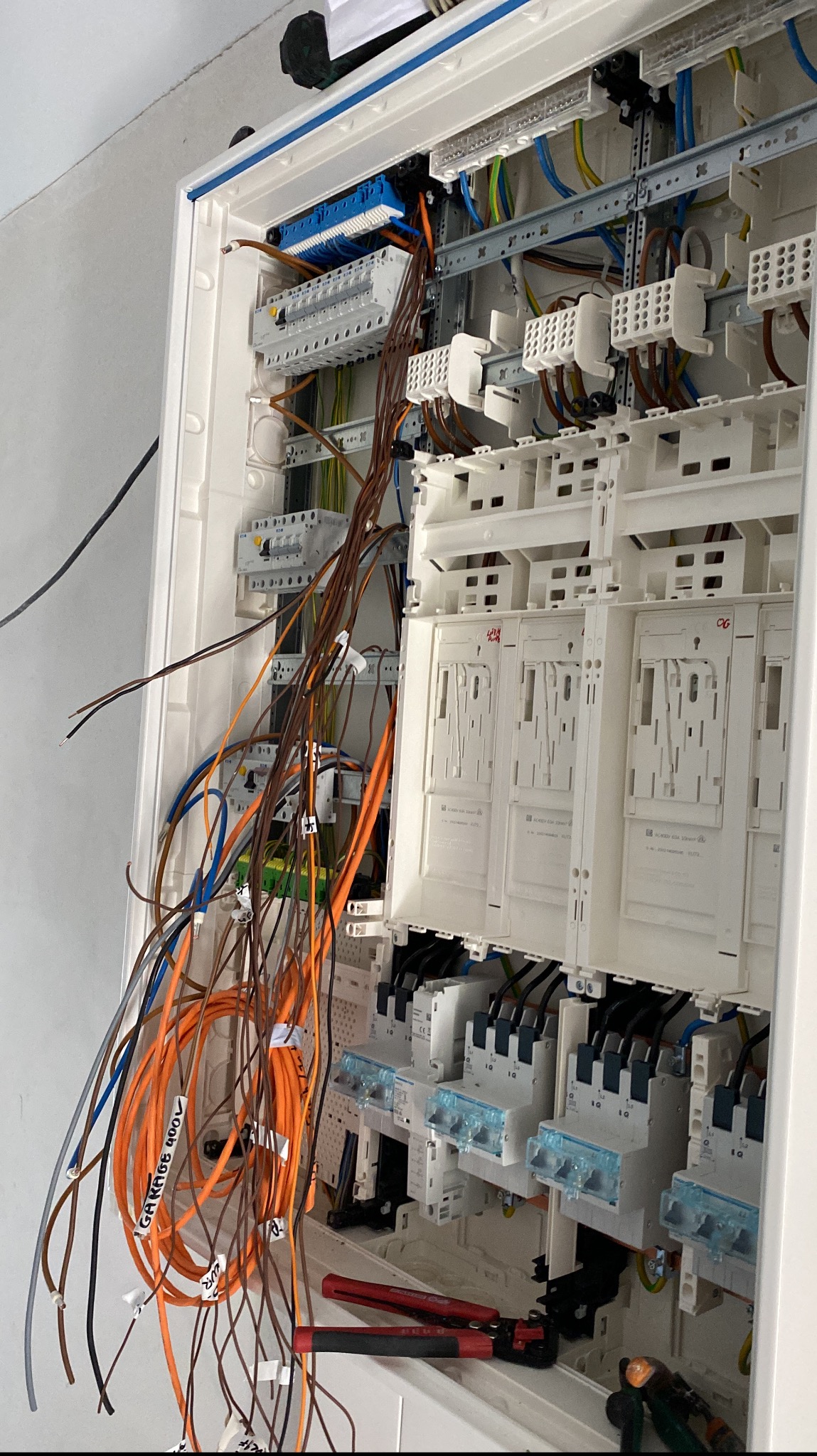 Rozdzielnia elektryczna w trakcie montażu: widoczne przewody z oznaczeniami, bezpieczniki i narzędzia (szczypce). Biała obudowa, szare ściany w tle.
