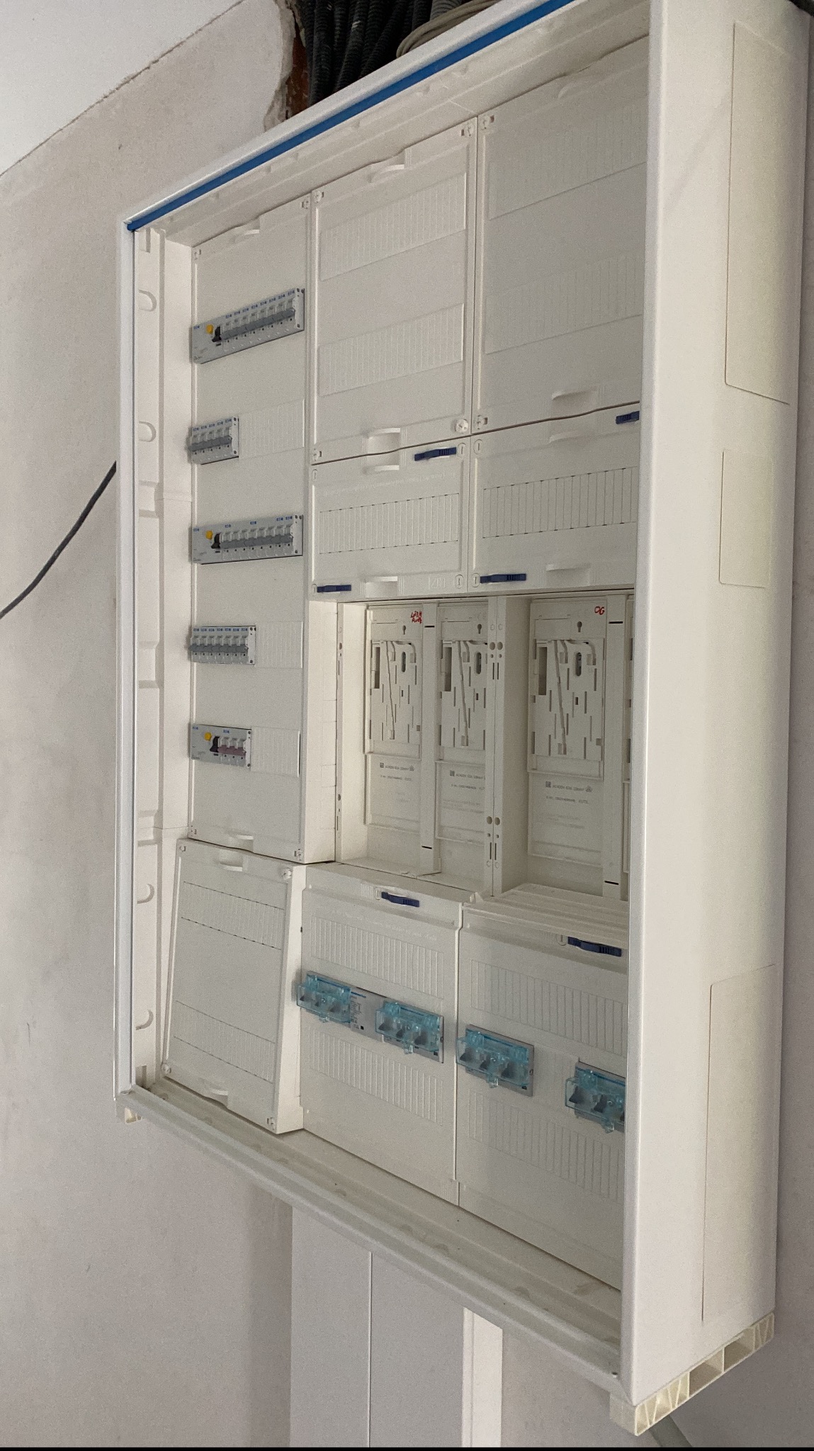 Otwarta, biała skrzynka rozdzielcza z widocznymi bezpiecznikami i elementami instalacji elektrycznej, zamontowana na ścianie wewnątrz budynku.