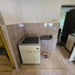 Beżowy piec CO z rurą odprowadzającą spaliny, obok ułożone drewno na podłodze z beżowych płytek. W tle fragment kuchni i wejście do innego pomieszczenia.