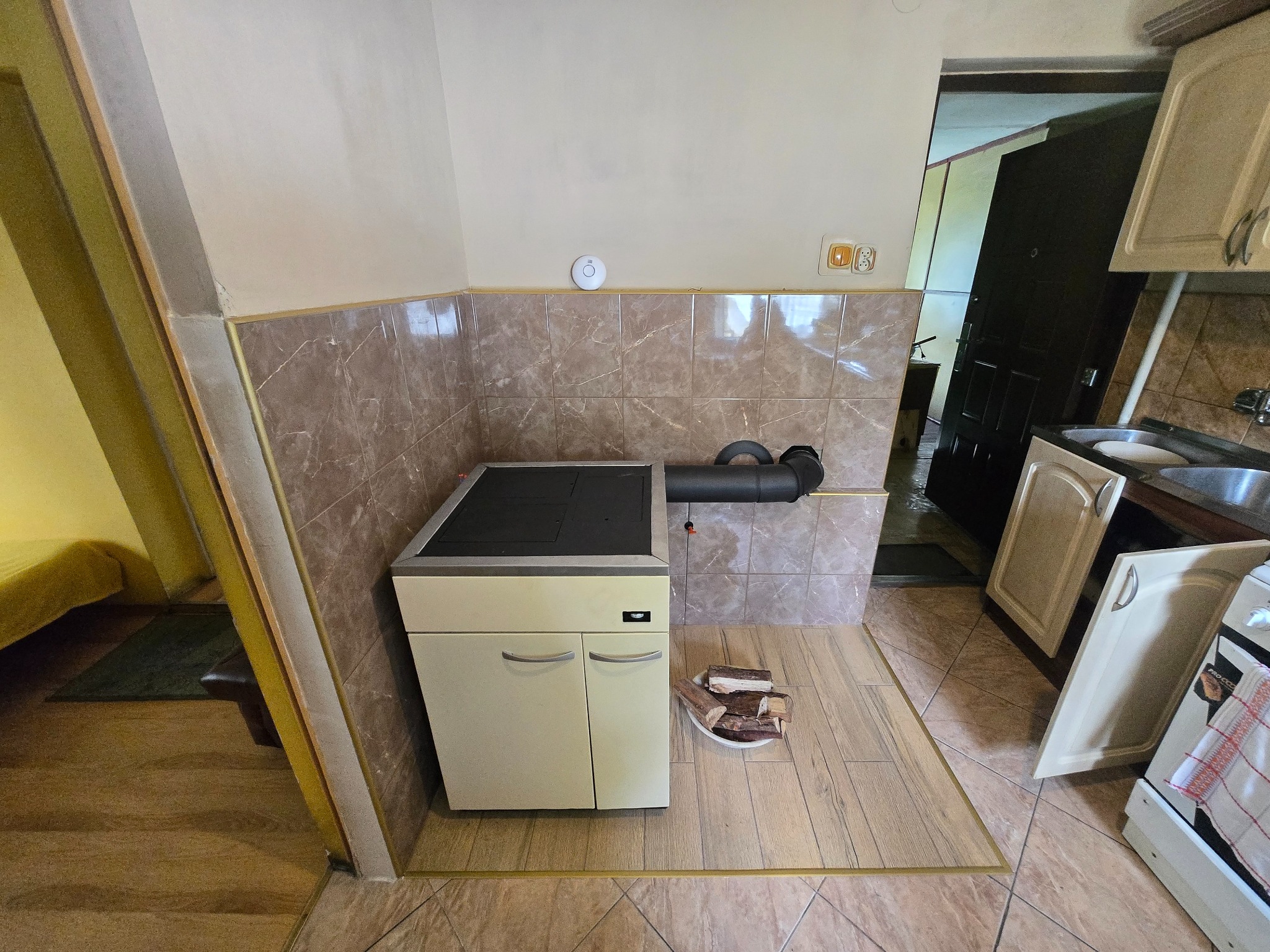 Beżowy piec CO z rurą odprowadzającą spaliny, obok ułożone drewno na podłodze z beżowych płytek. W tle fragment kuchni i wejście do innego pomieszczenia.