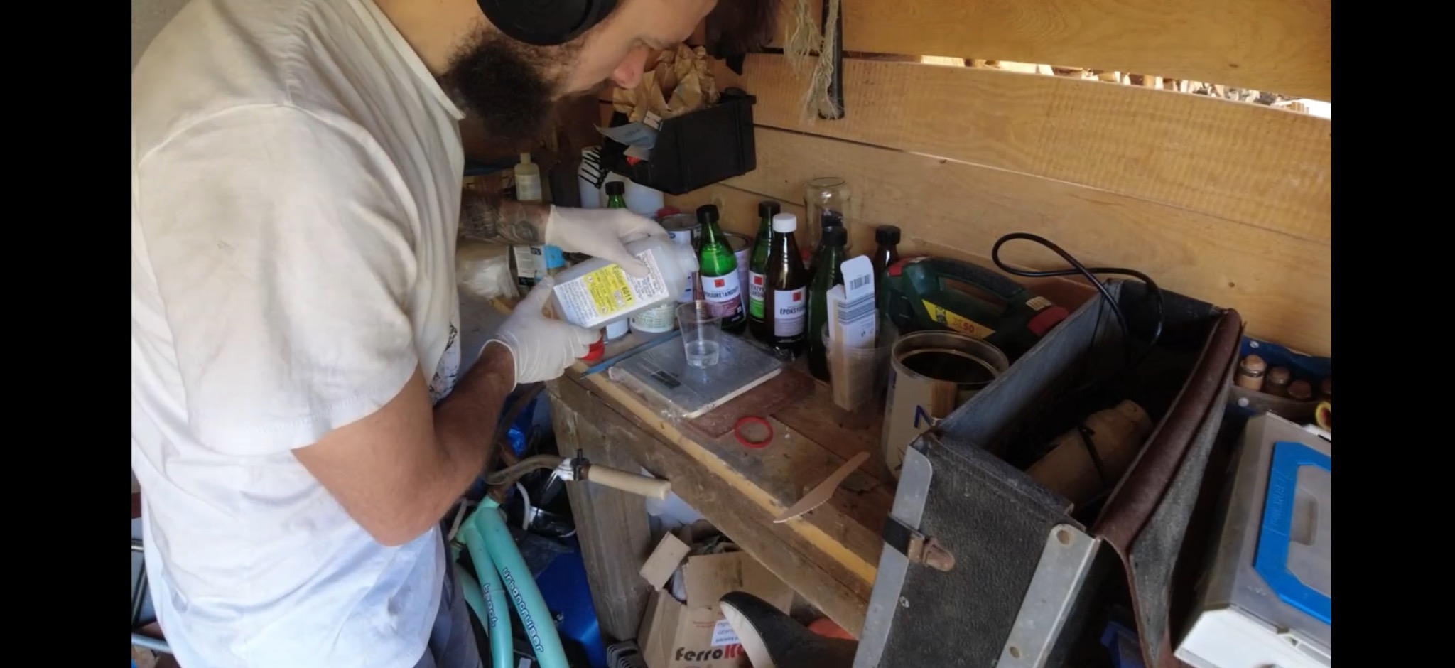 Osoba w rękawicach wlewa płyn z butelki do pojemnika na wadze. W tle butelki, puszki, narzędzia i drewniana ściana. Nieuporządkowane miejsce pracy.