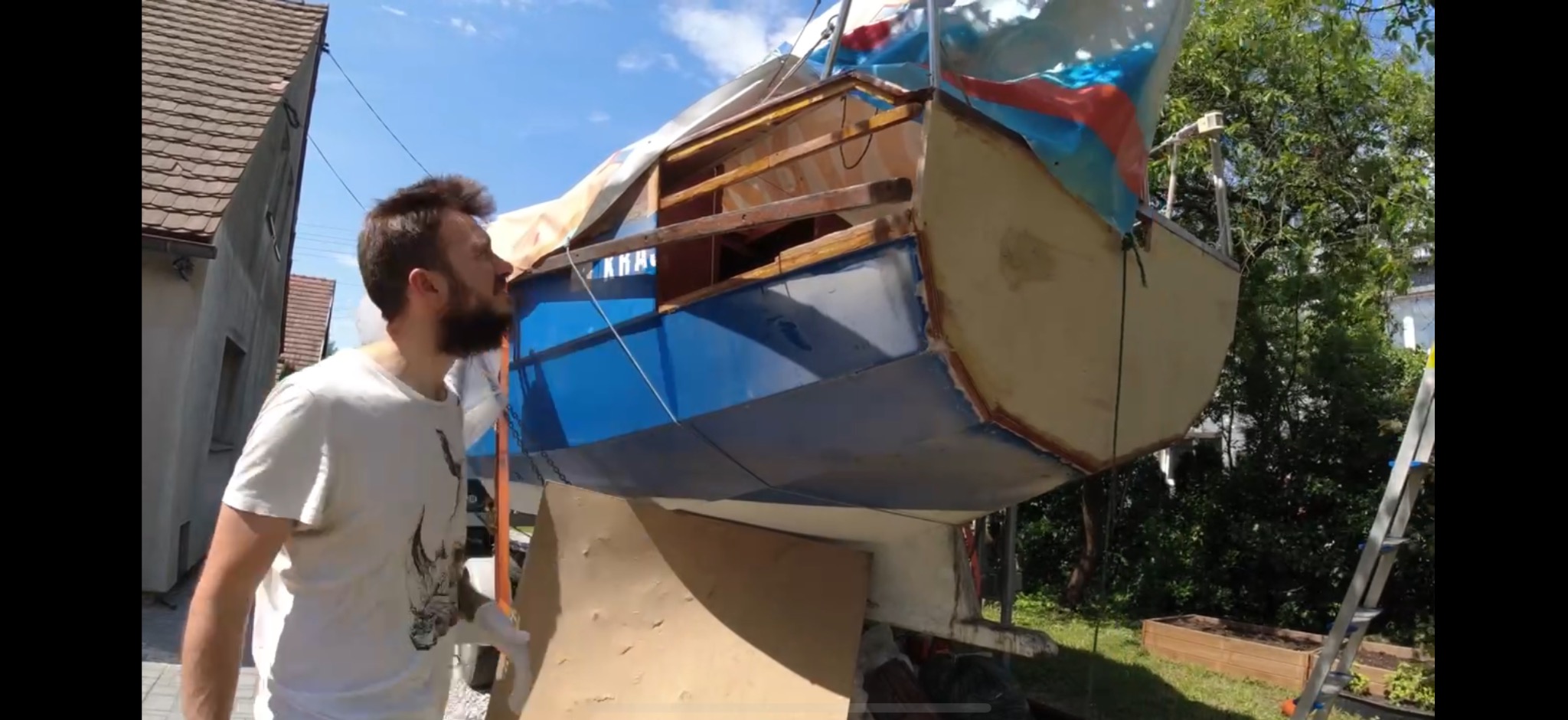 Mężczyzna ogląda odnowioną, niebiesko-białą łódź, stojącą na zewnątrz. W tle dom i zieleń. Widoczne ślady prac renowacyjnych.