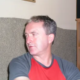 Mężczyzna w czerwono-szarej koszulce, patrzy w lewo. W tle ściana z fakturą i drewniany regał z telefonem. Ujęcie z lewej strony.