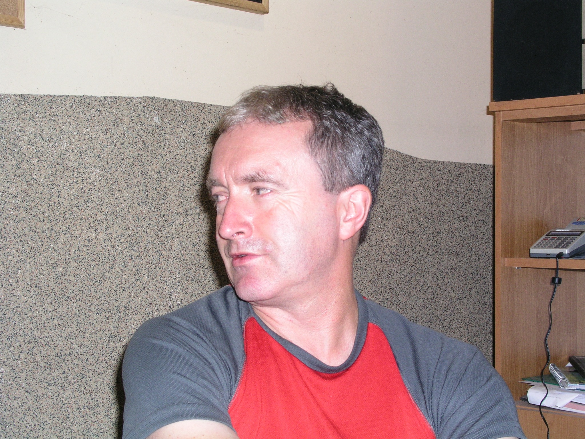 Mężczyzna w czerwono-szarej koszulce, patrzy w lewo. W tle ściana z fakturą i drewniany regał z telefonem. Ujęcie z lewej strony.