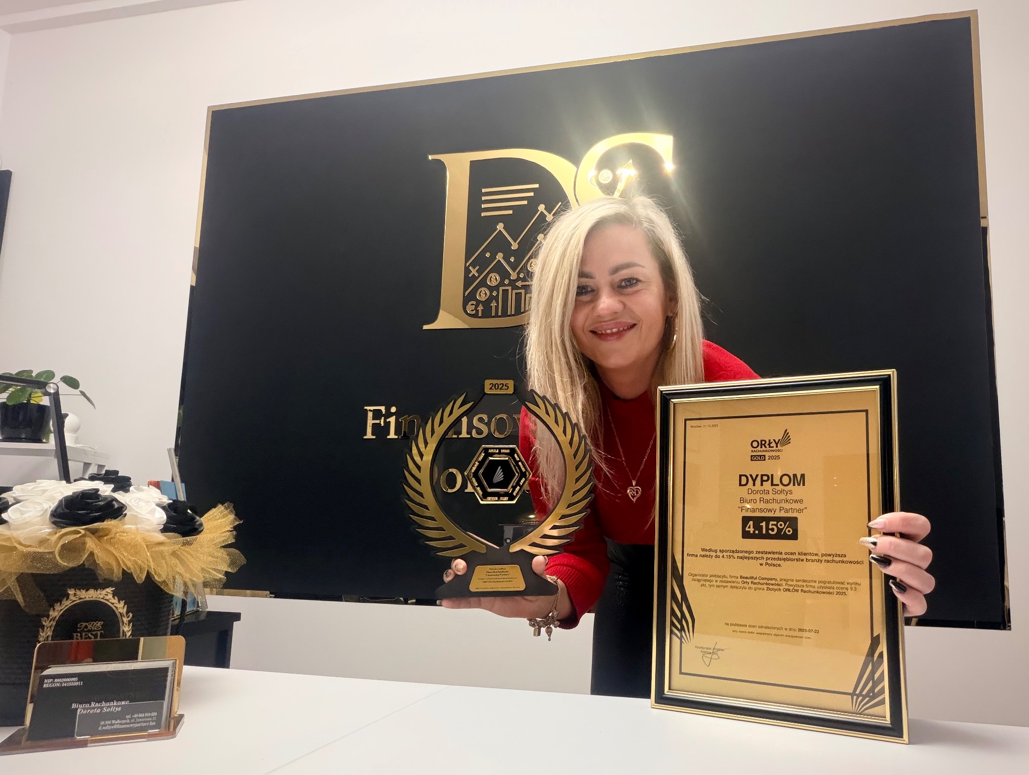 Kobieta z blond włosami prezentuje nagrody: statuetkę i dyplom 'Orły Rachunkowości Gold 2025' na tle logo firmy finansowej. Na biurku wizytówki i dekoracje.
