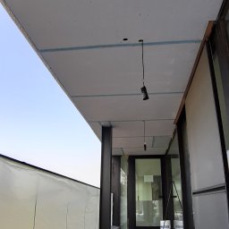 Marcin Szarek - Montaż płyt karton-gips na suficie z widocznymi profilami, przygotowane podłoże i wiszące lampy. Widoczne narzędzia pracy i zabezpieczenia.